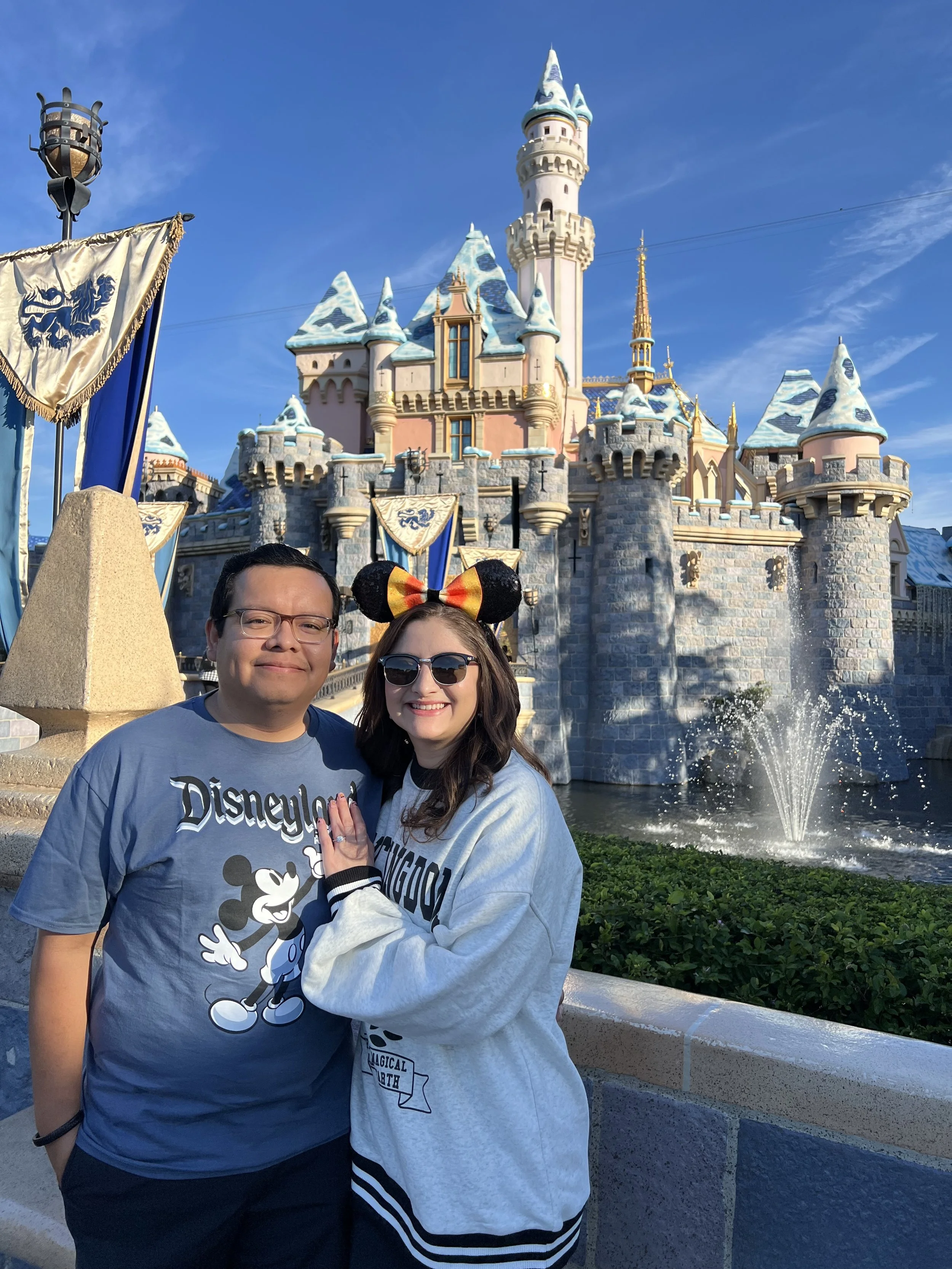 The owner and his wife are smiling at the camera in front of Sleeping Beauty Castle at Disneyland with banners, a fountain, and blue sky in the background.