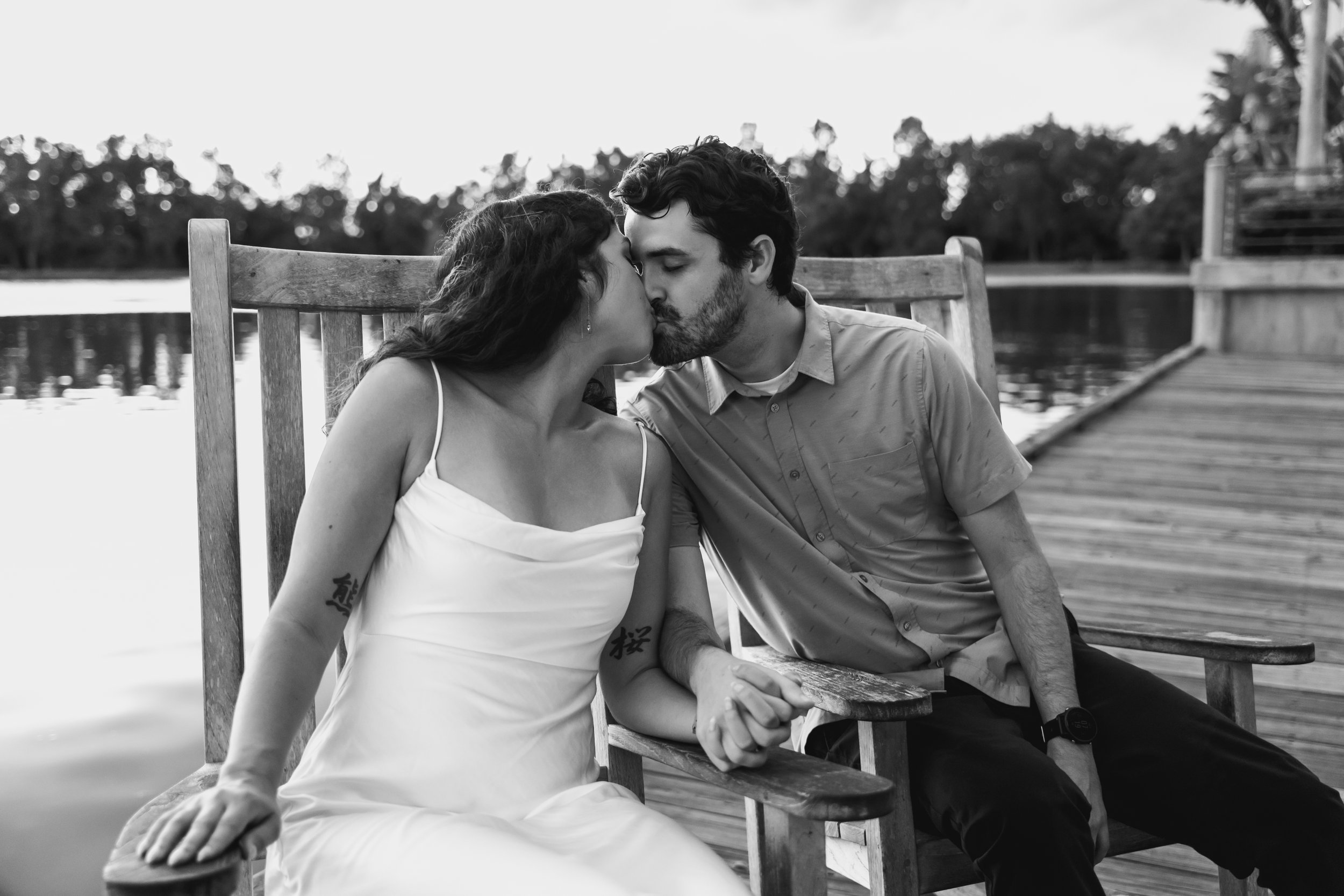 A black and white photo of a couple sitting on a park bench, holding hands and kissing by a lake with trees in the background.