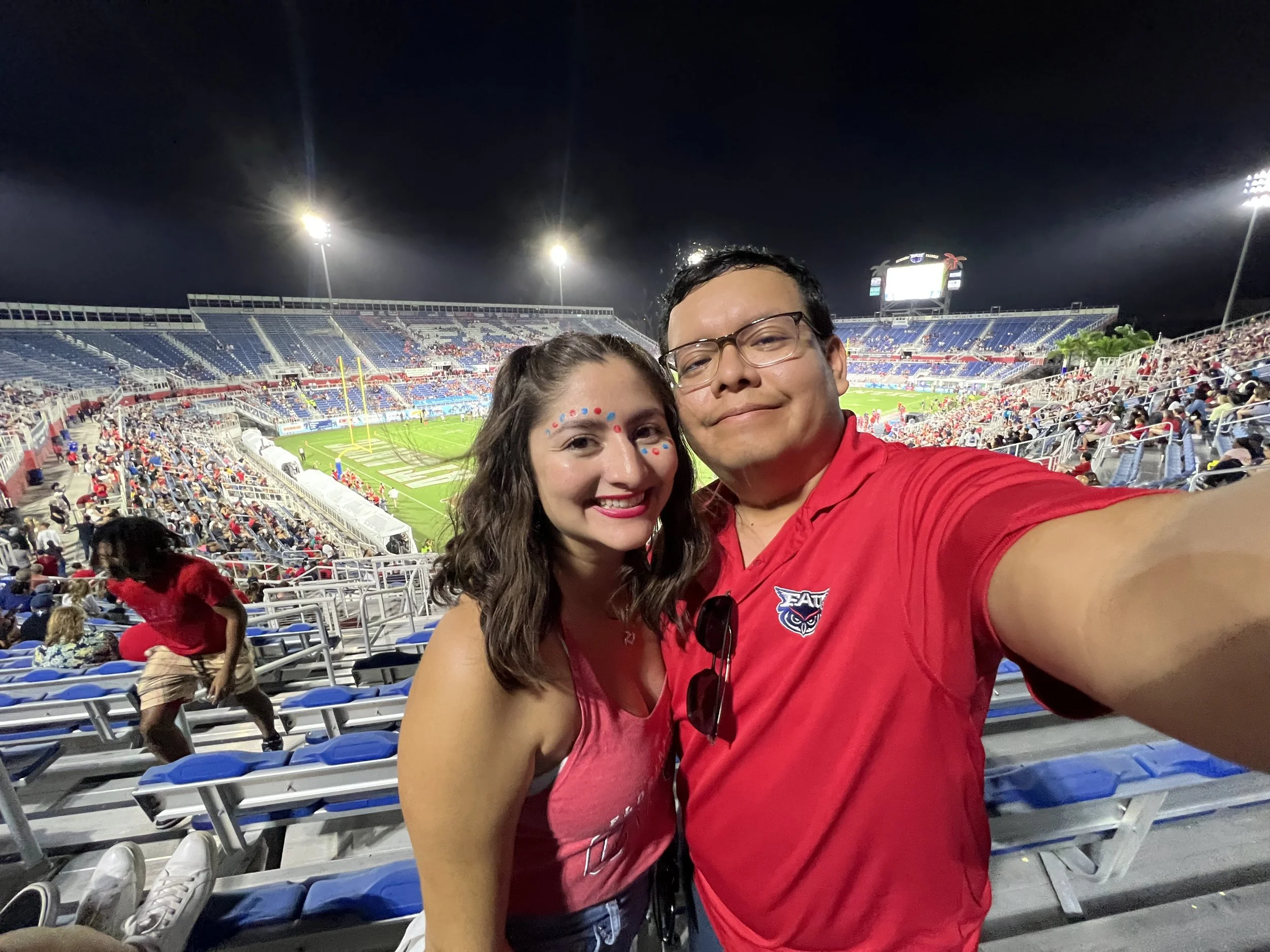 The owner and his wife are taking a selfie at a crowded stadium during a night game, with field and empty bleacher seats visible in the background.