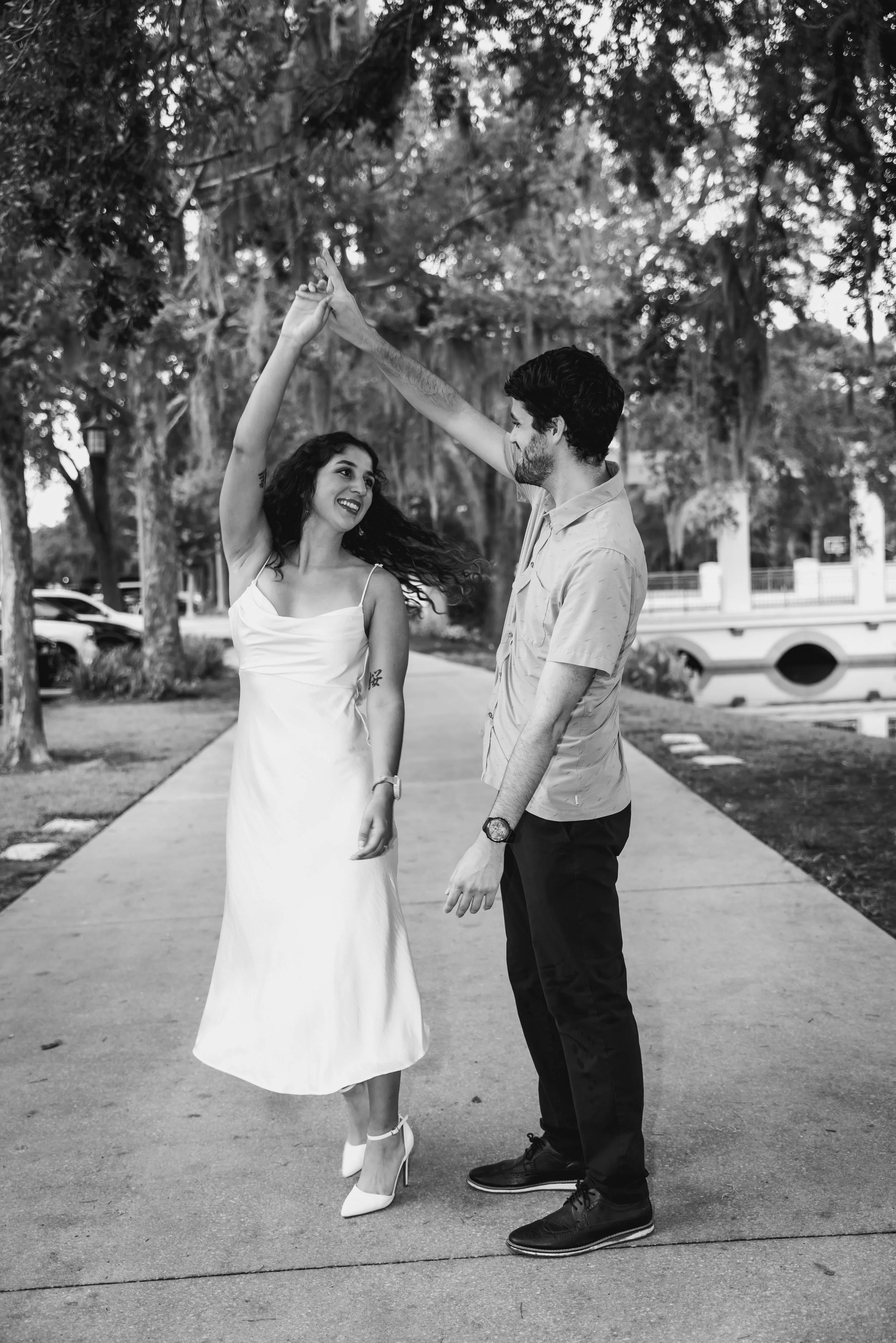 A couple dancing outdoors on a sidewalk, with trees and a bench in the background. The woman wears a white dress and heels, and the man wears a short-sleeve shirt and dark pants. They are looking at each other and smiling as they dance.
