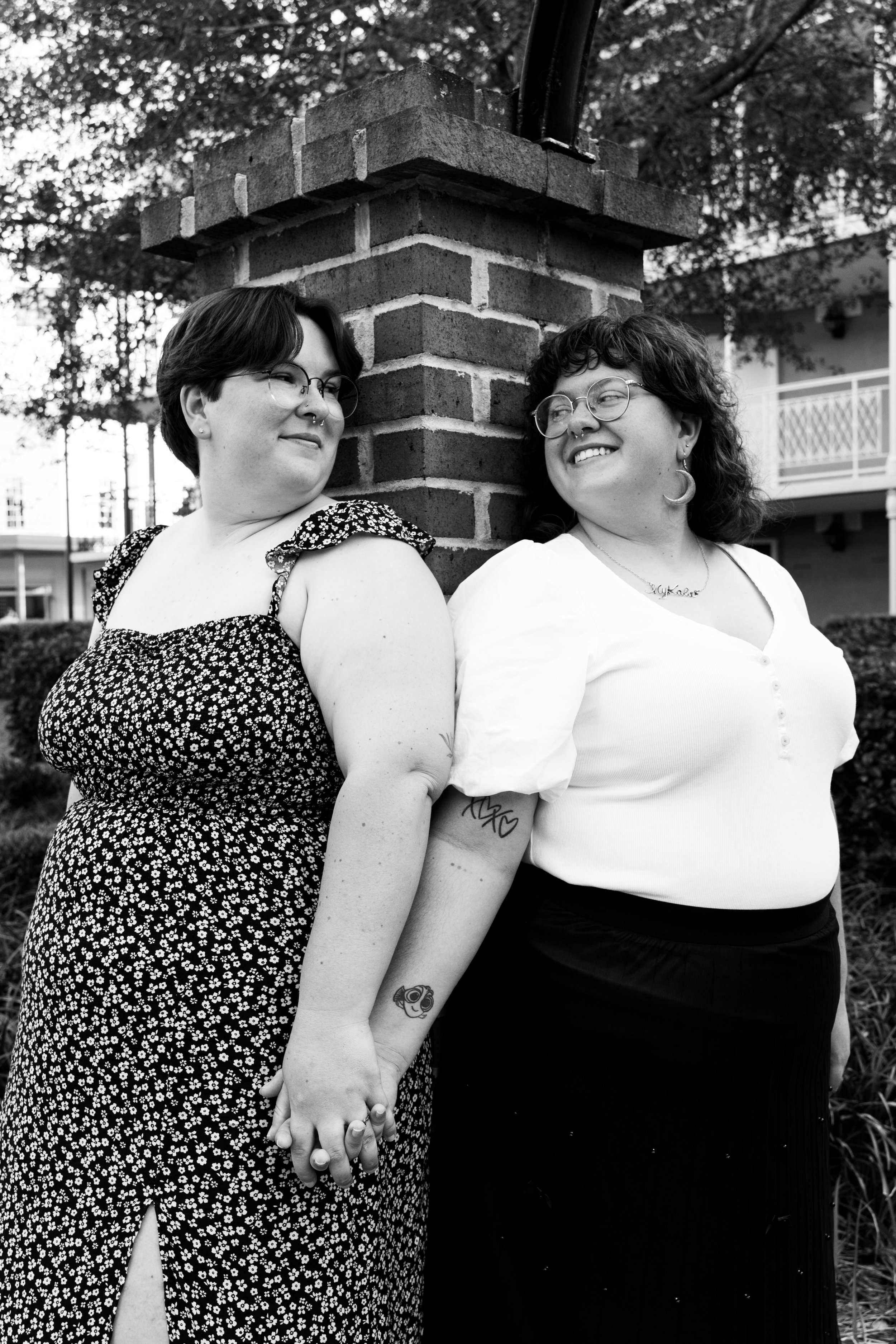 Two women standing face to face, holding hands, leaning against a brick post outdoors. One woman has short dark hair, glasses, and wears a floral dress. The other has curly hair, glasses, and wears a white top and a black skirt.