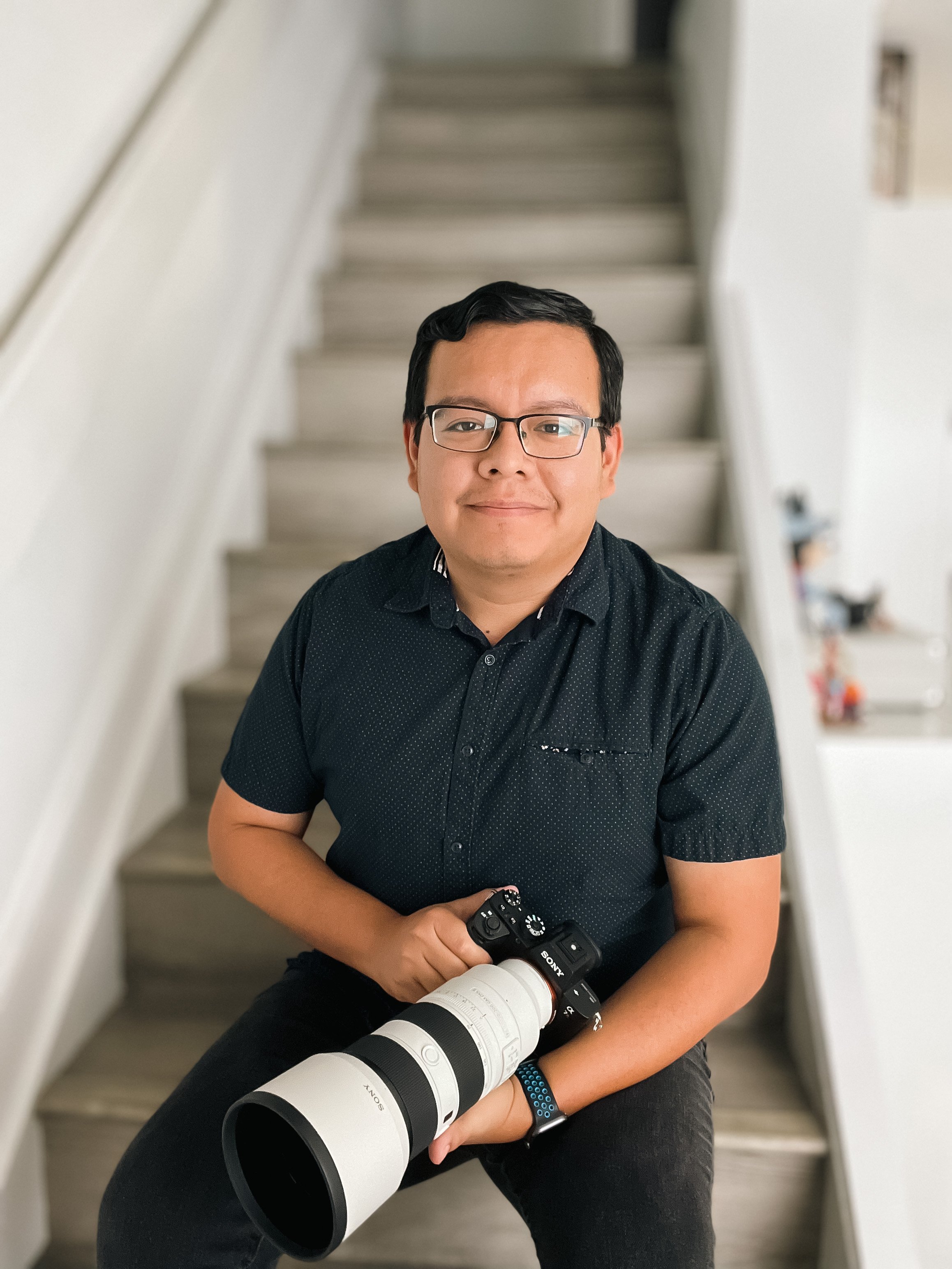 A man, the owner of AAR Photography, wearing glasses and a black shirt, sitting on stairs holding a professional camera with a large telephoto lens.