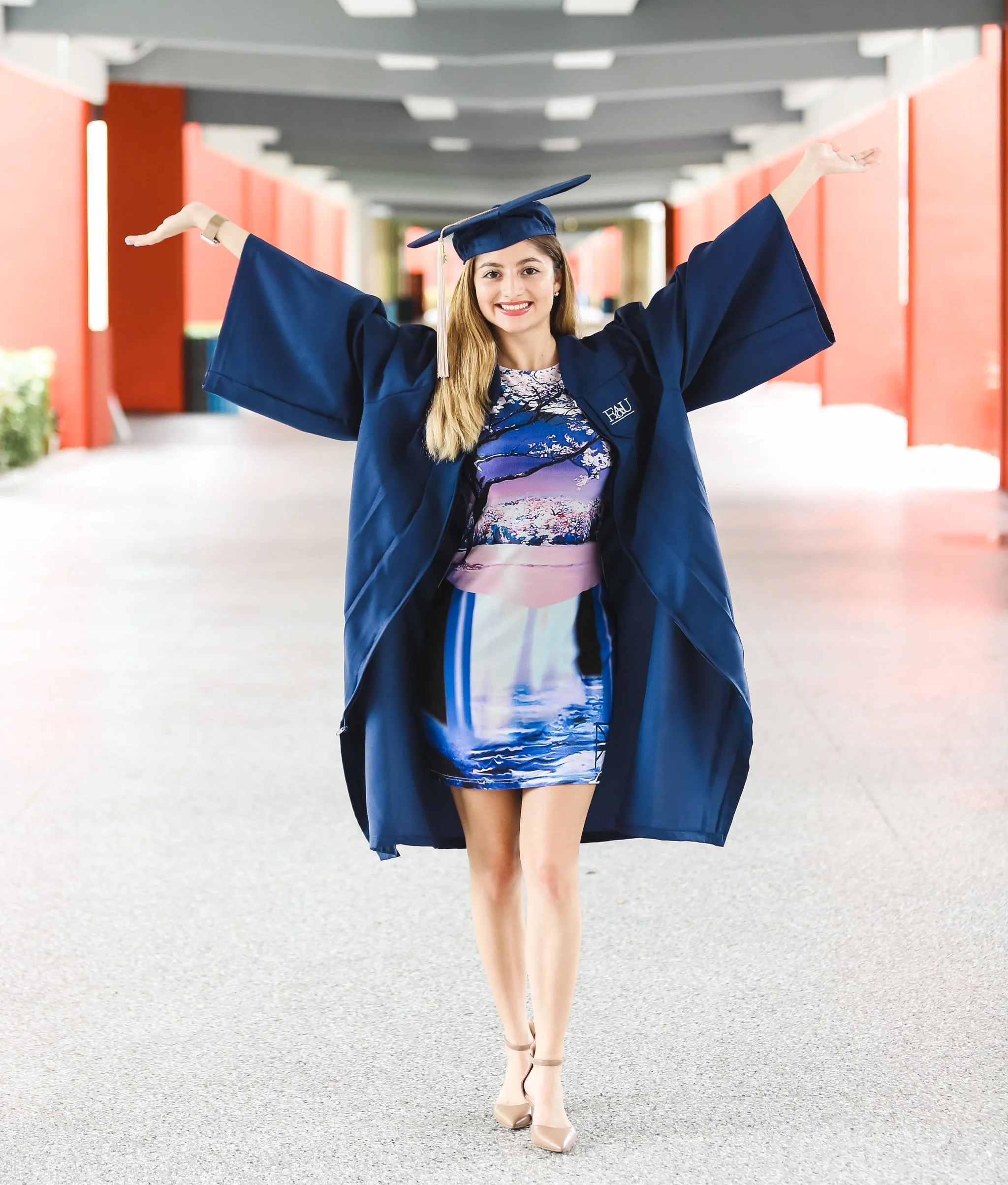 A young woman in a blue graduation gown and cap standing in an outdoor corridor with red walls, smiling, with her arms raised in celebration.