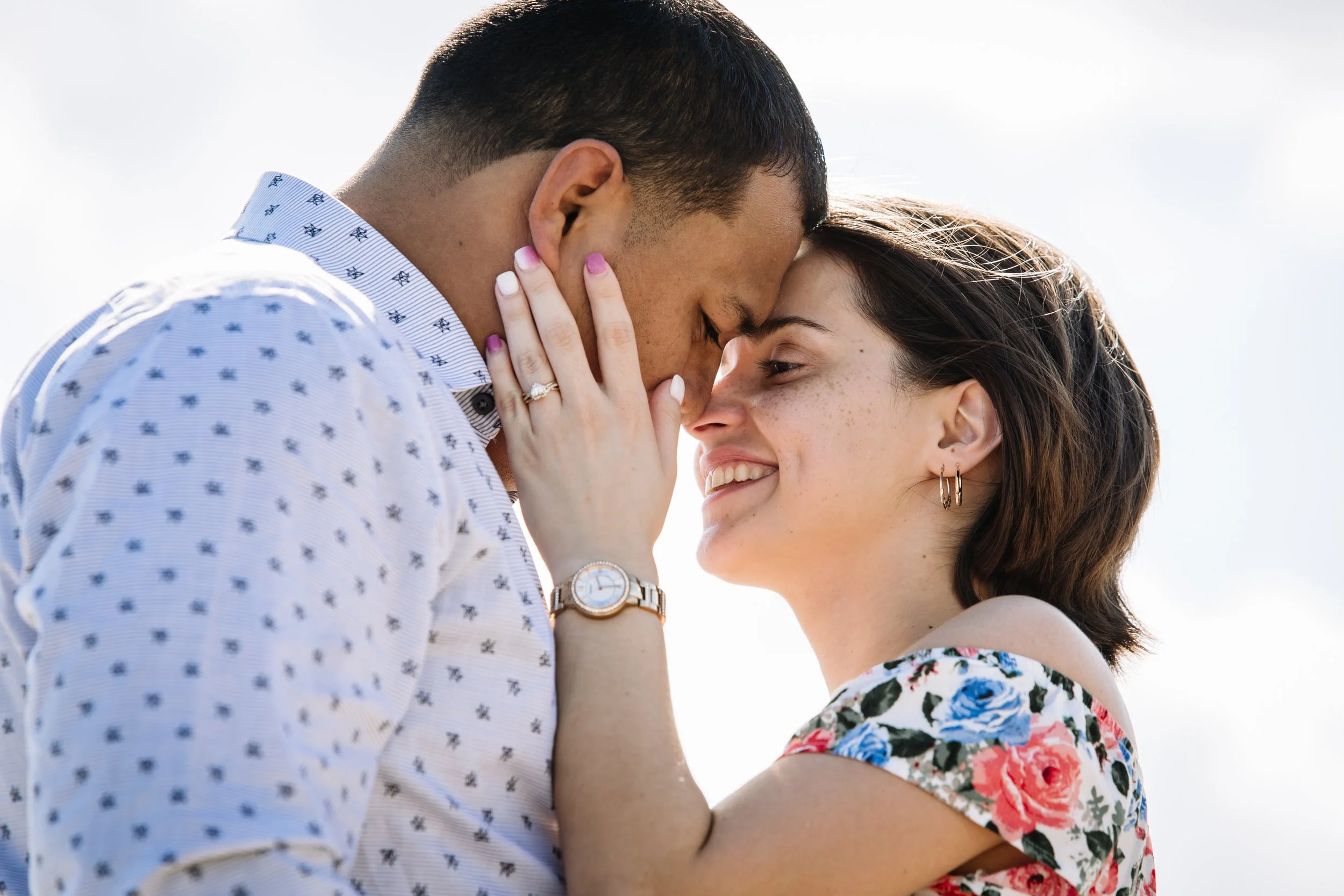 A couple is touching foreheads and smiling. The man is dressed in a white shirt with a small pattern and is wearing a watch. The woman is wearing earrings and a floral dress. The background is bright, and they are outdoors.