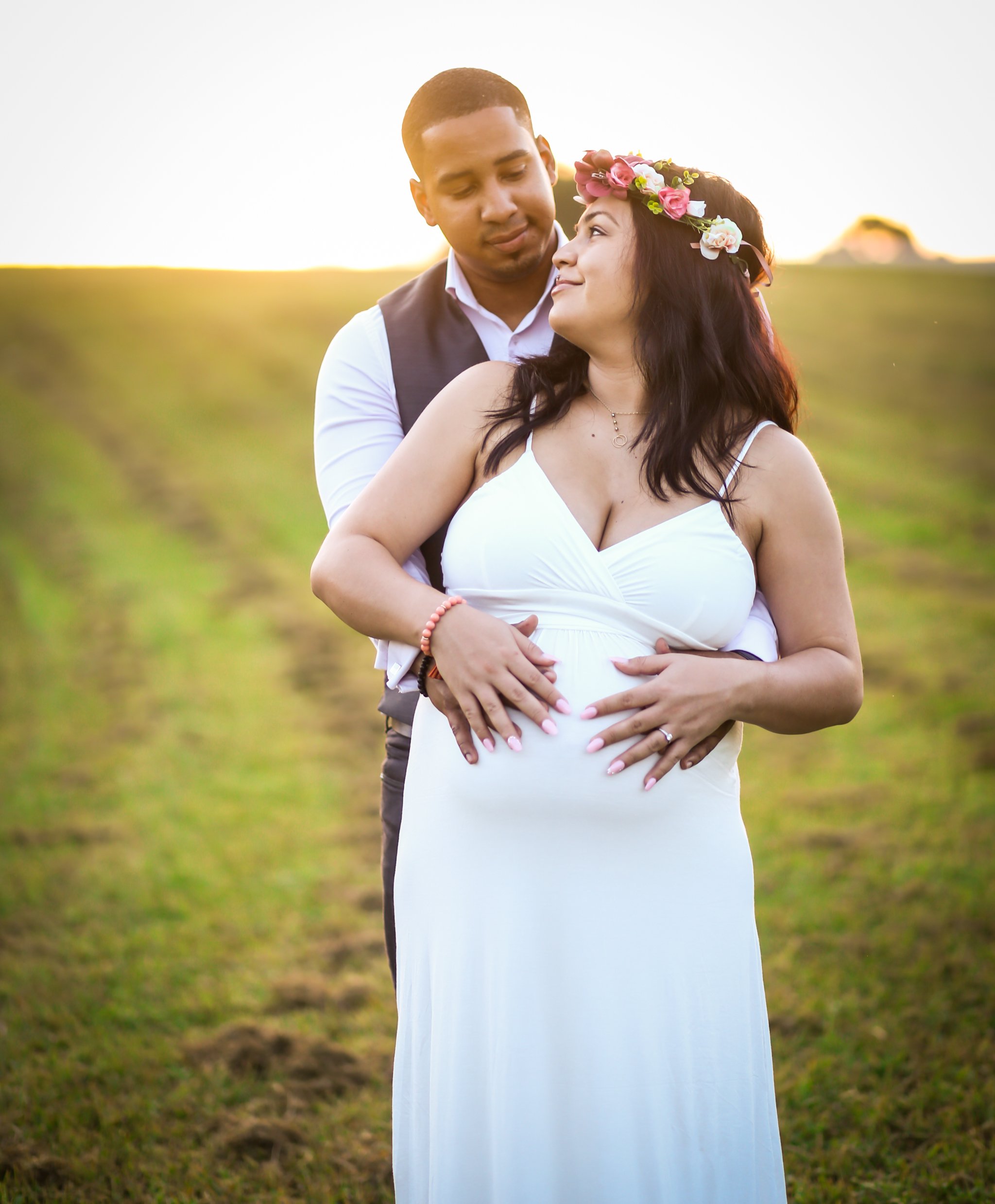 A man and woman embrace outdoors during sunset, woman wearing a white dress and floral crown, man in vest and white shirt, green field in background.
