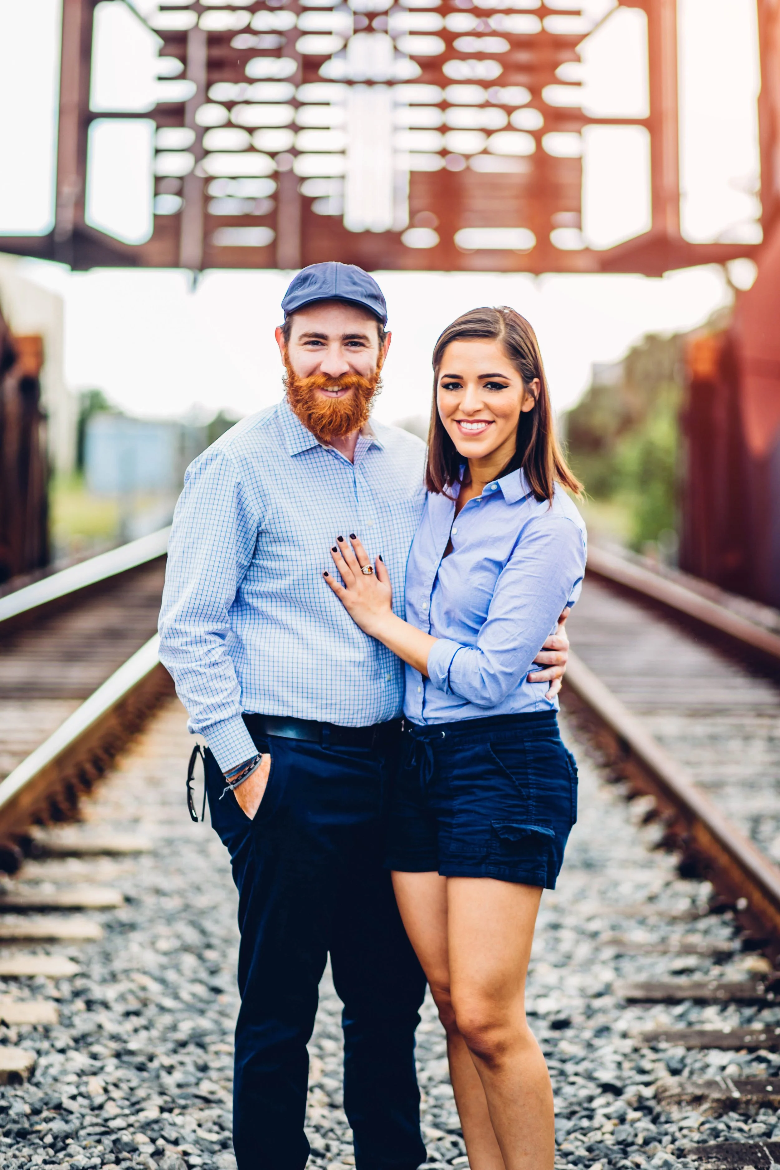 A young man with a red beard, wearing a blue baseball cap and blue checkered shirt, and a young woman with brown hair, wearing a blue shirt and black shorts, standing together on a railway track under a bridge, smiling at the camera.