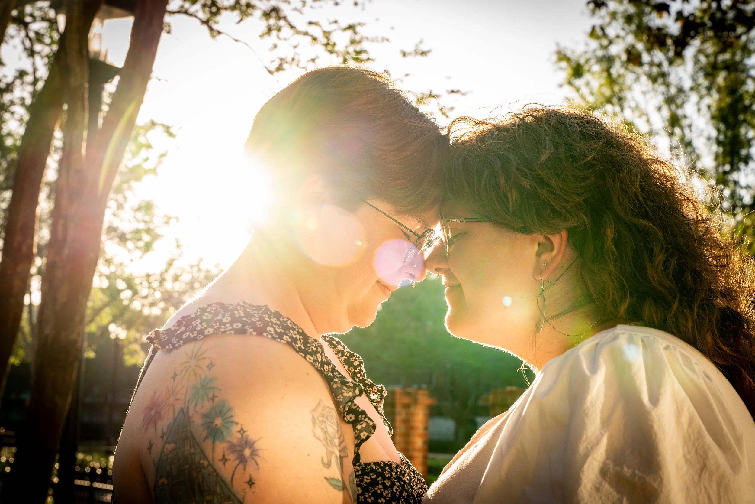Two women with glasses and curly hair, face to face outdoors in sunlight, touching foreheads, with trees in the background.