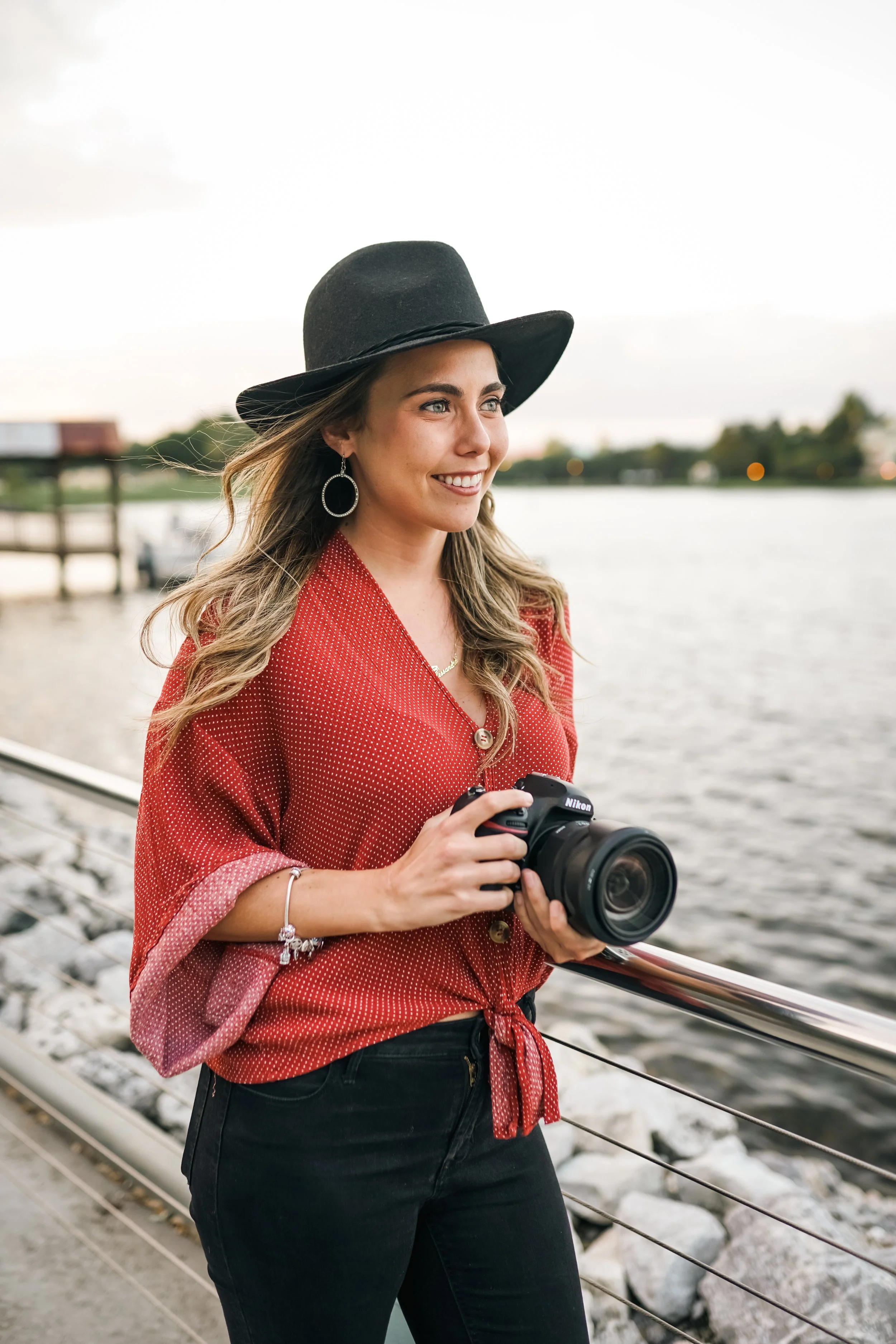 A young woman with long wavy hair, wearing a black wide-brimmed hat, hoop earrings, a red blouse with white polka dots, and black pants, is holding a Nikon camera while standing near a body of water on a cloudy day.