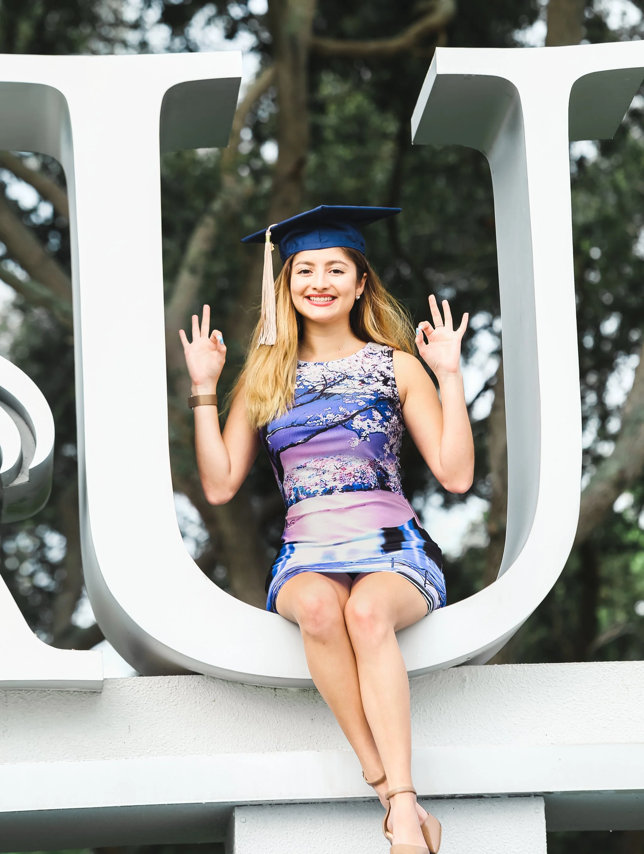 A young woman wearing a graduation cap and a colorful sleeveless dress sitting on a large white sculpture that resembles an open book. She is smiling and flashing the peace sign with both hands.