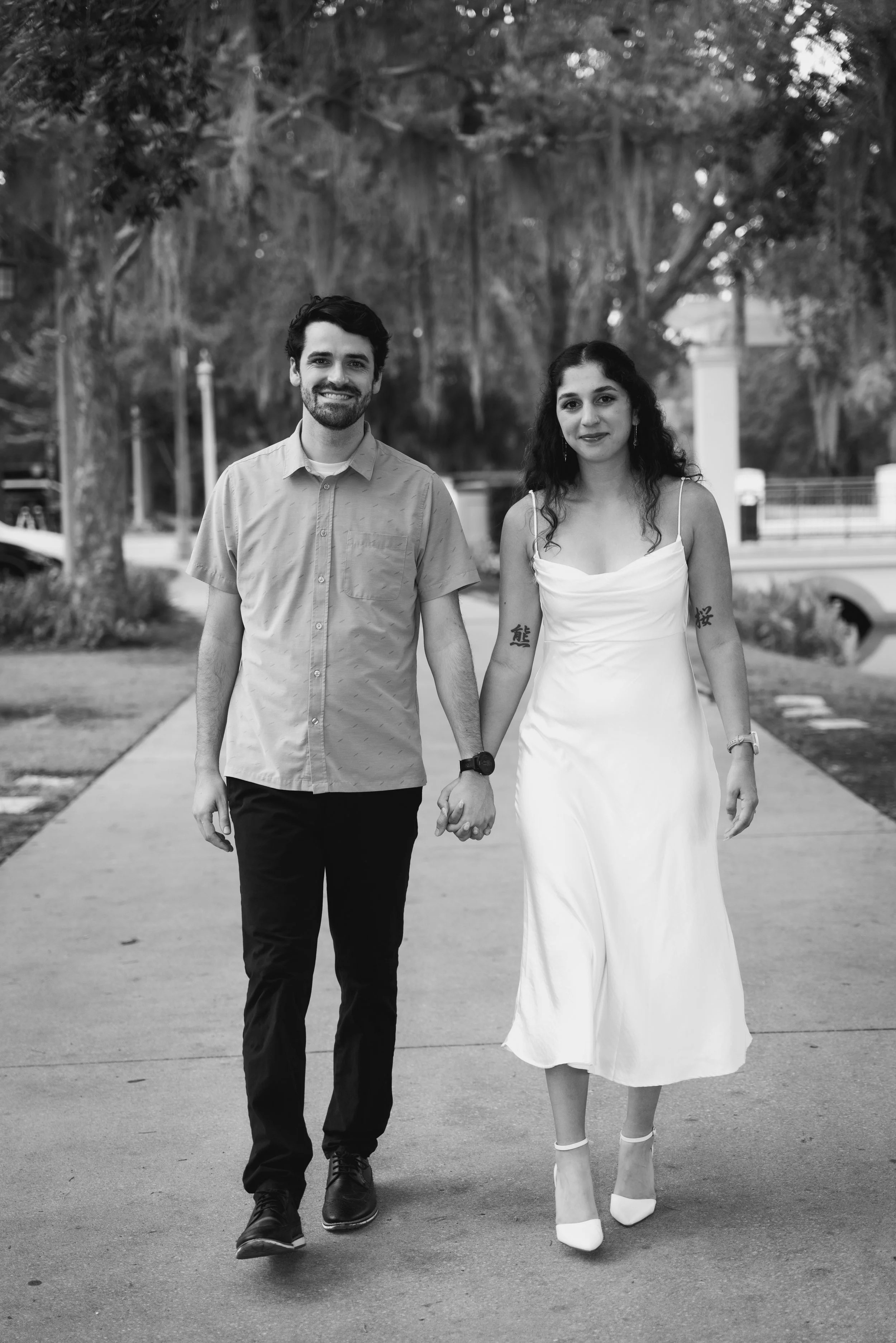 A smiling man and woman holding hands and walking on a sidewalk with trees in the background. The woman is wearing a white dress and heels, and the man is wearing a button-up shirt and pants.