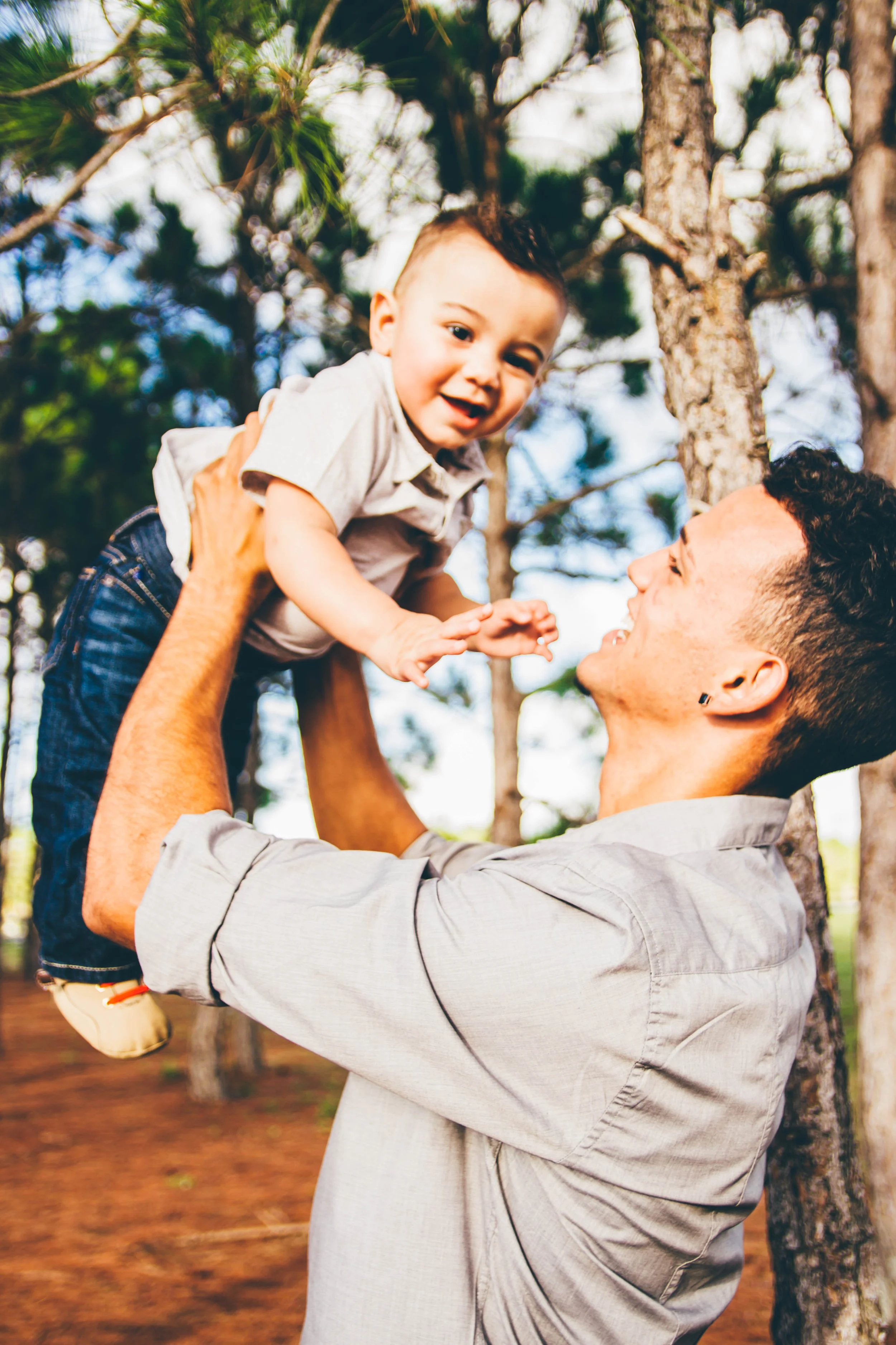 A man lifting a young boy in a park with trees in the background, both smiling and enjoying the moment.