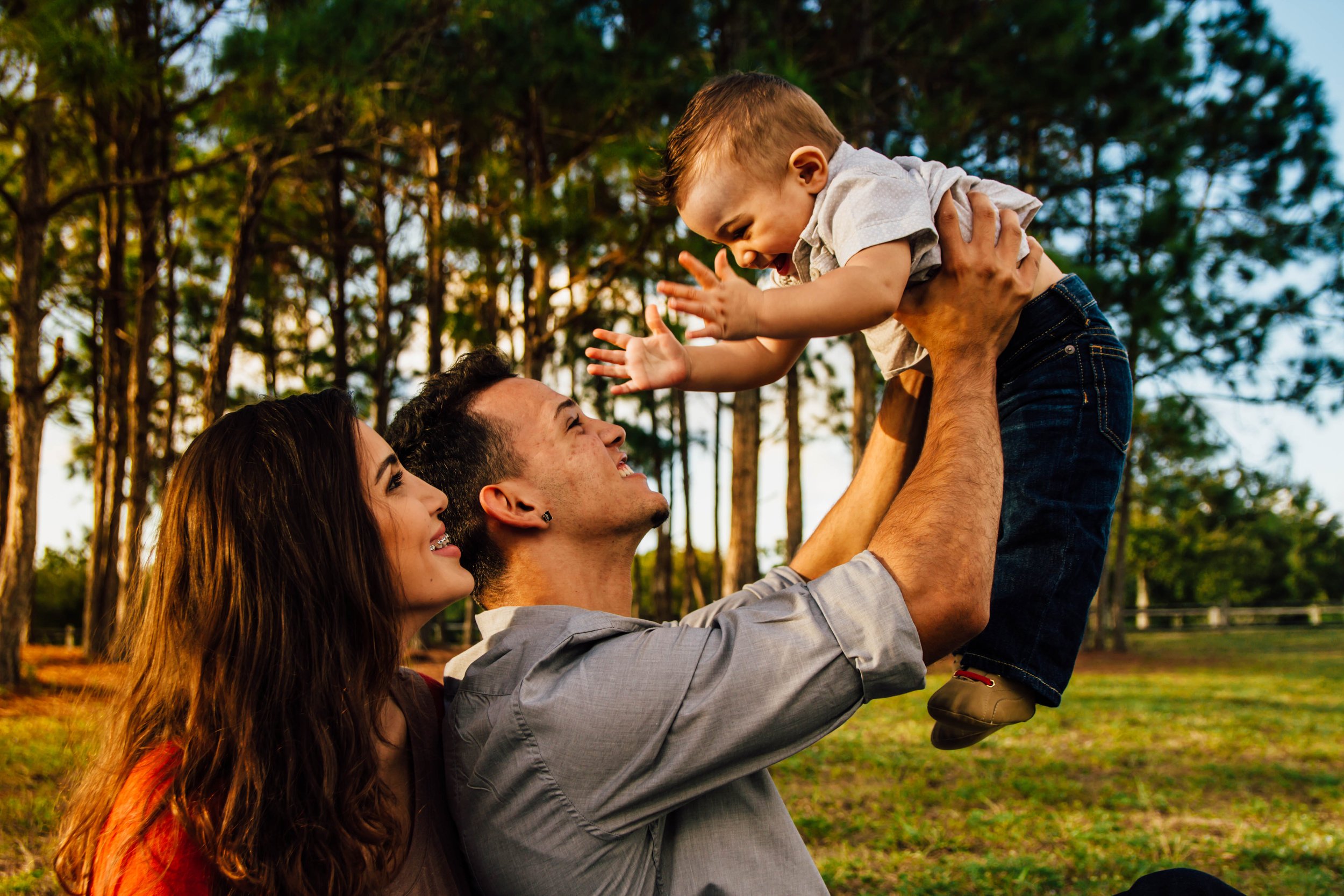 A family of three outdoors, with two adults and a child, enjoying a sunny day in a wooded park. The adults are holding the child up in the air, and they all look happy and smiling.