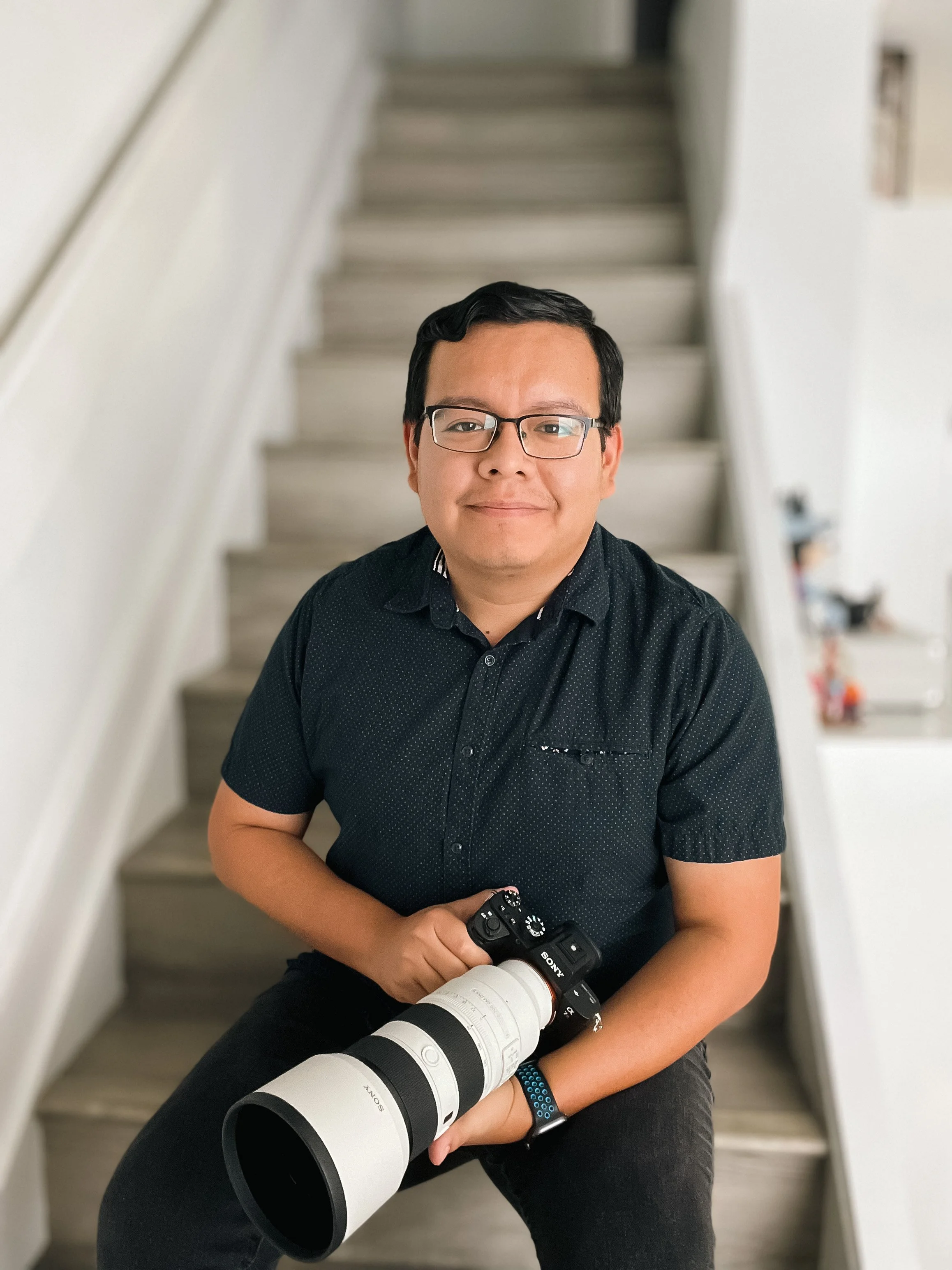Portrait of a man, owner of AAR Photography, with glasses, dark hair, wearing a black shirt, sitting on stairs inside a house, holding a professional camera with a large telephoto lens.