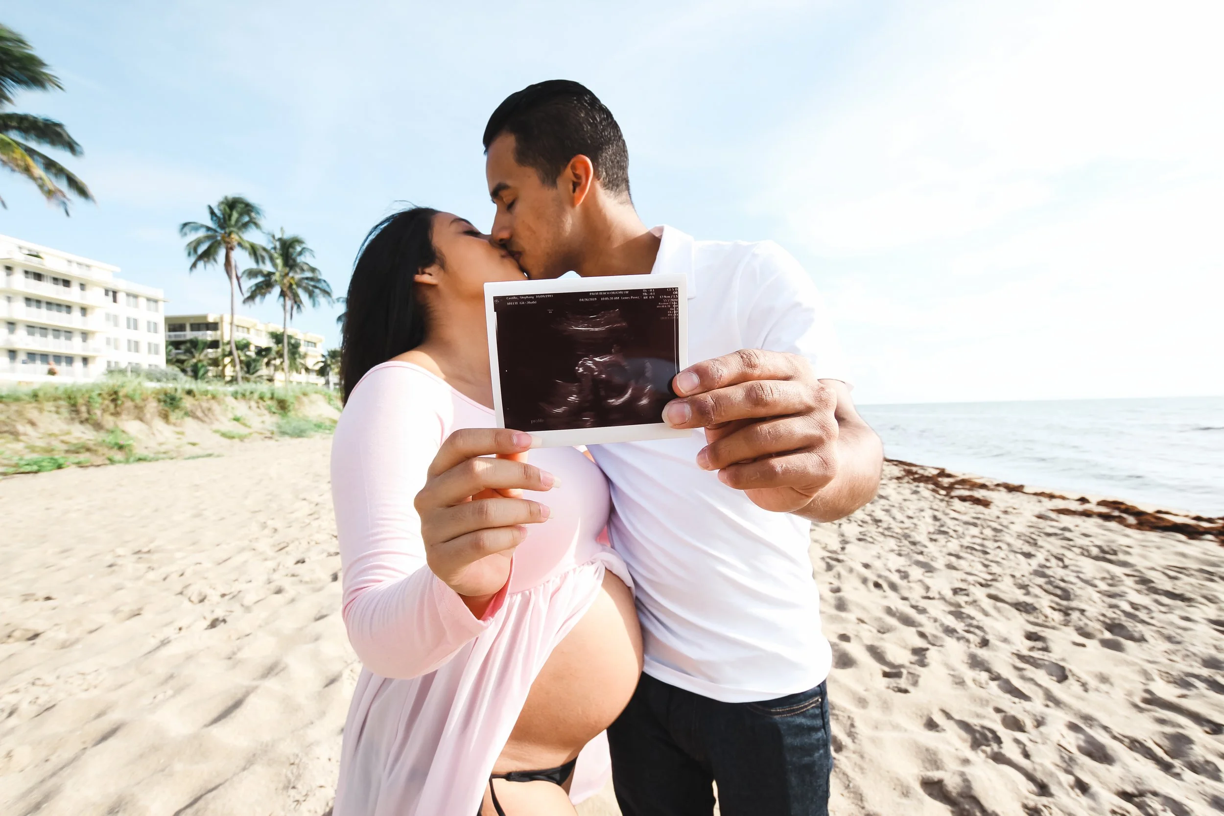 A couple passionately kissing on the beach, holding an ultrasound image of a baby, with palm trees and beachside buildings in the background.