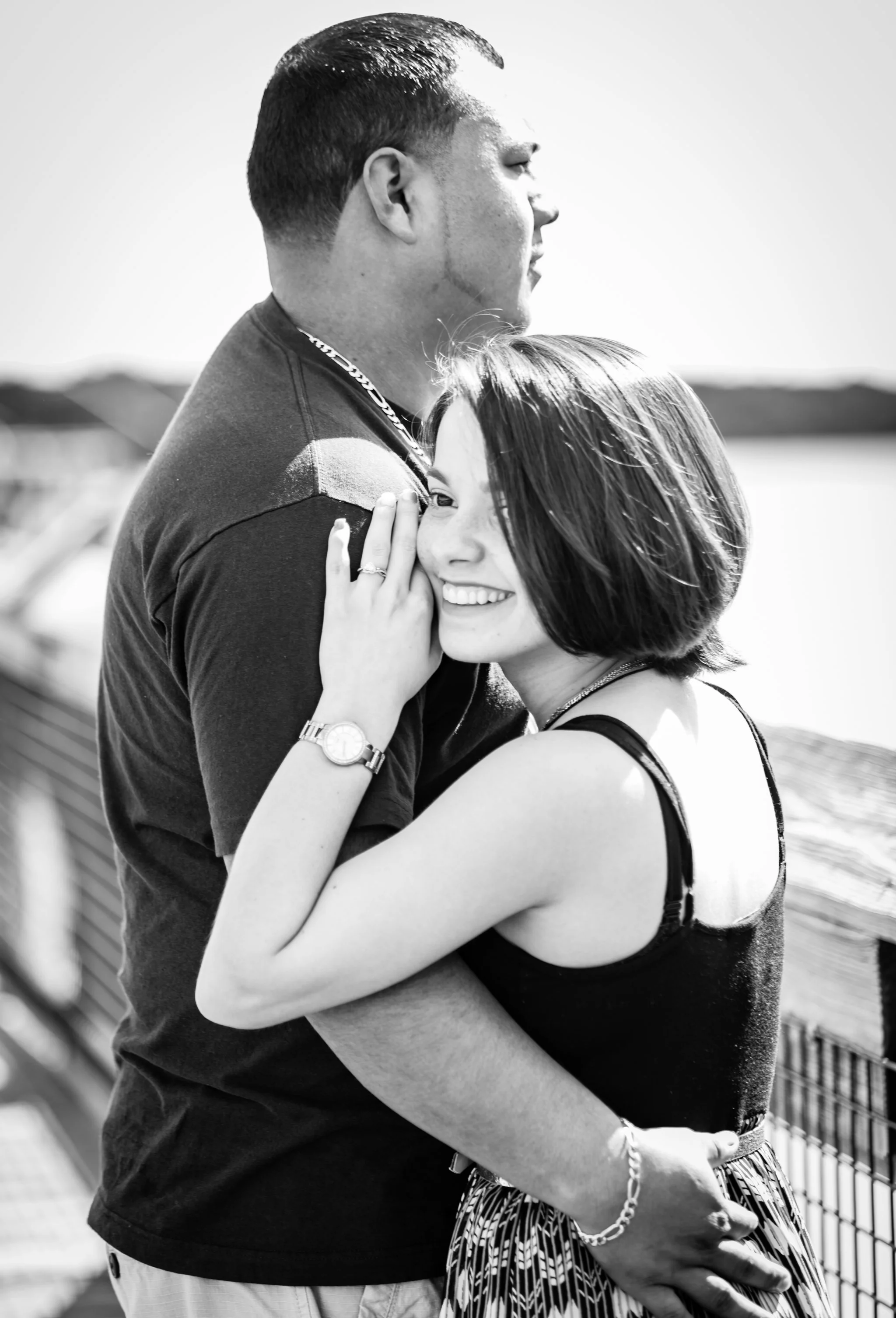 A black and white photo of a young couple hugging on a bridge. The man is seen in profile, wearing a dark t-shirt, and the woman is smiling with her eyes closed, wearing a sleeveless top and a patterned skirt.