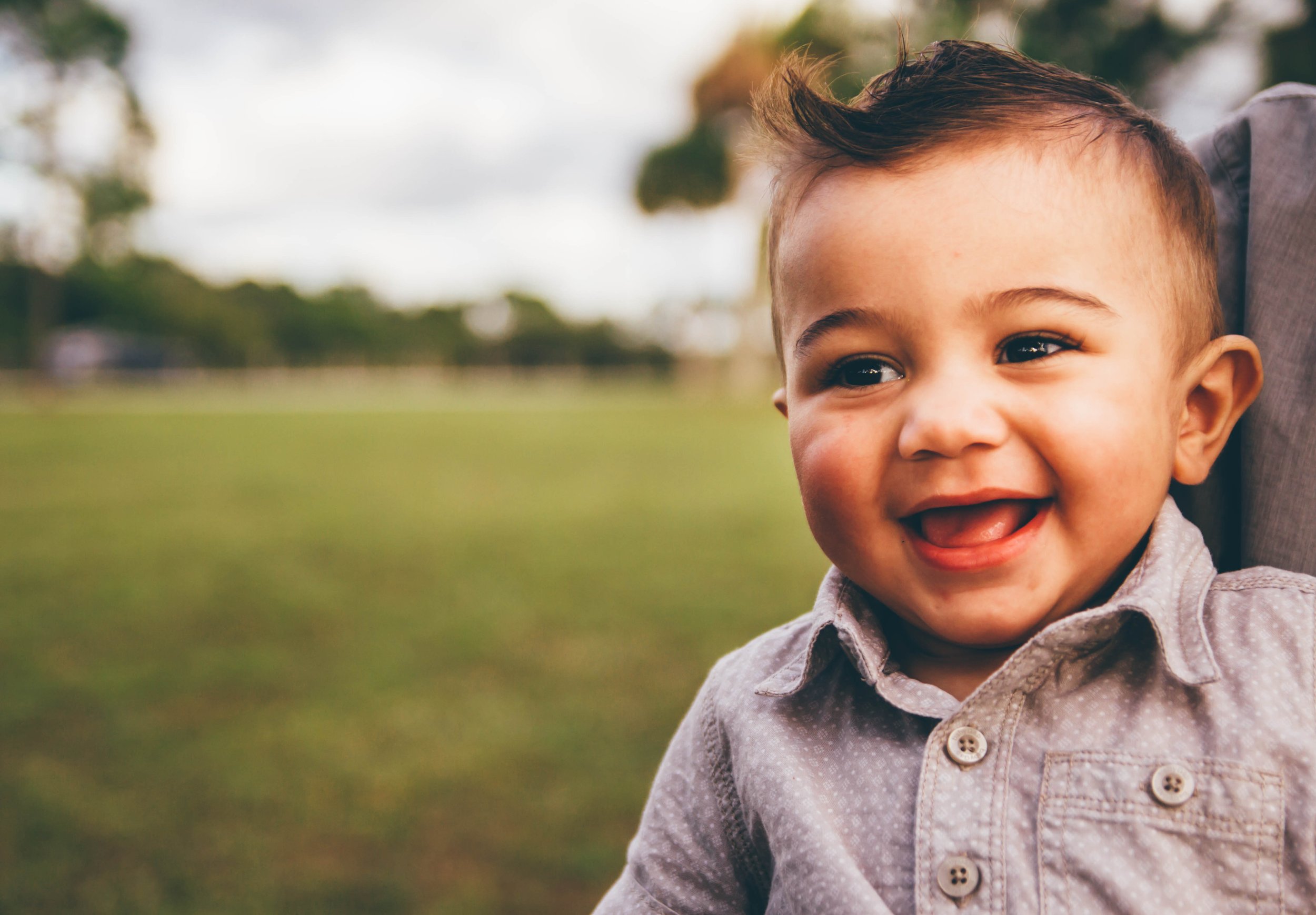 Close-up of a young child's face smiling outdoors, wearing a gray collared jacket, with blurred background of trees and grass.