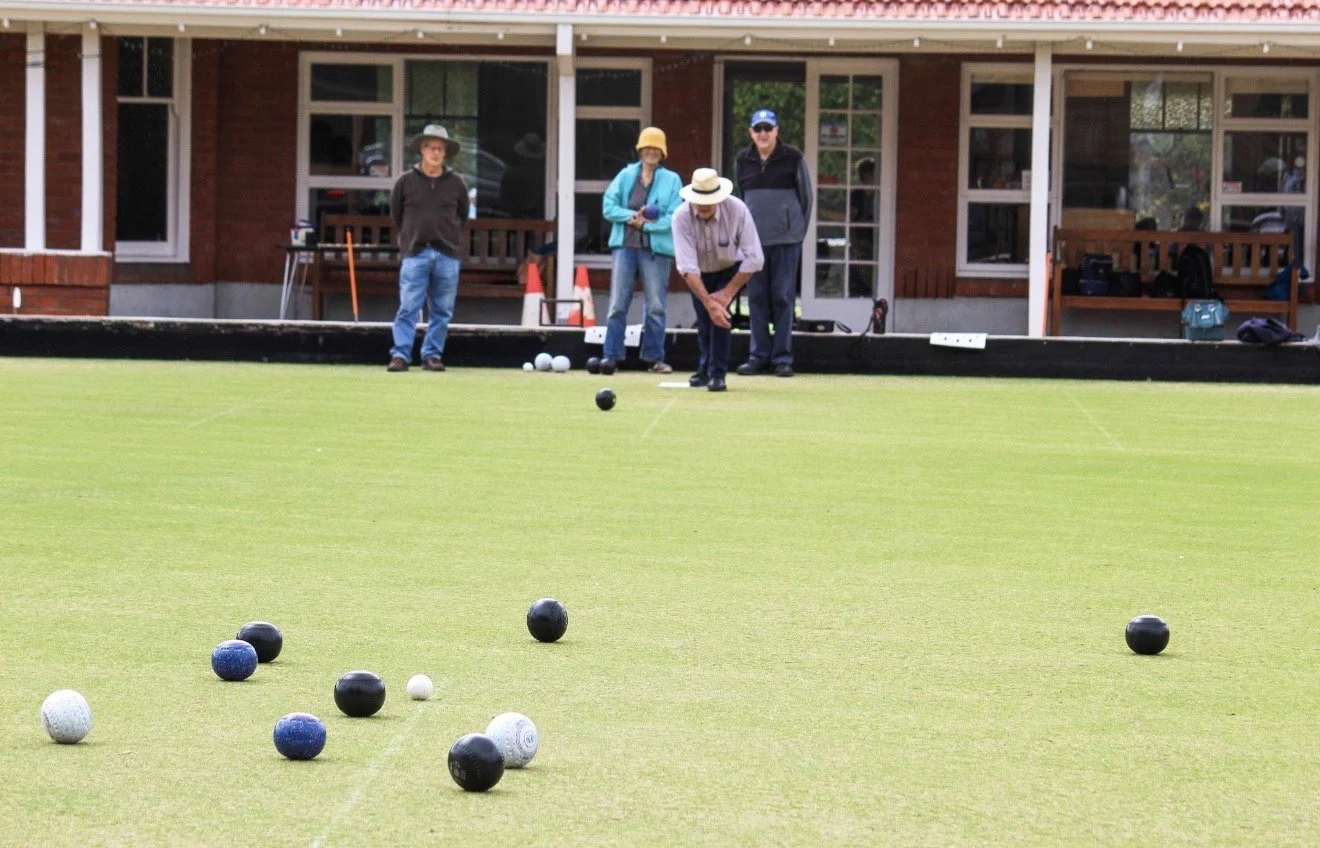 People playing lawn bowls on a green, with a brick house and porch in the background.