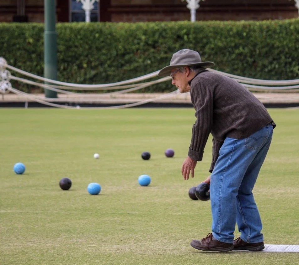 An elderly man wearing a gray hat, dark sweater, and jeans playing lawn bowls on a green field.