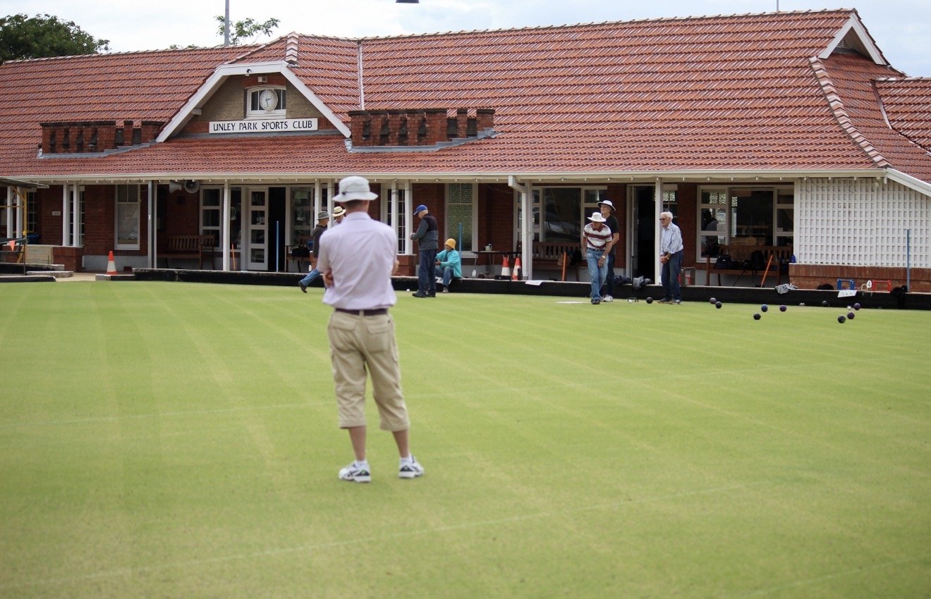 People playing lawn bowls on a green outside a brick clubhouse with a red tile roof, labeled 'Unley Park Sports Club'.