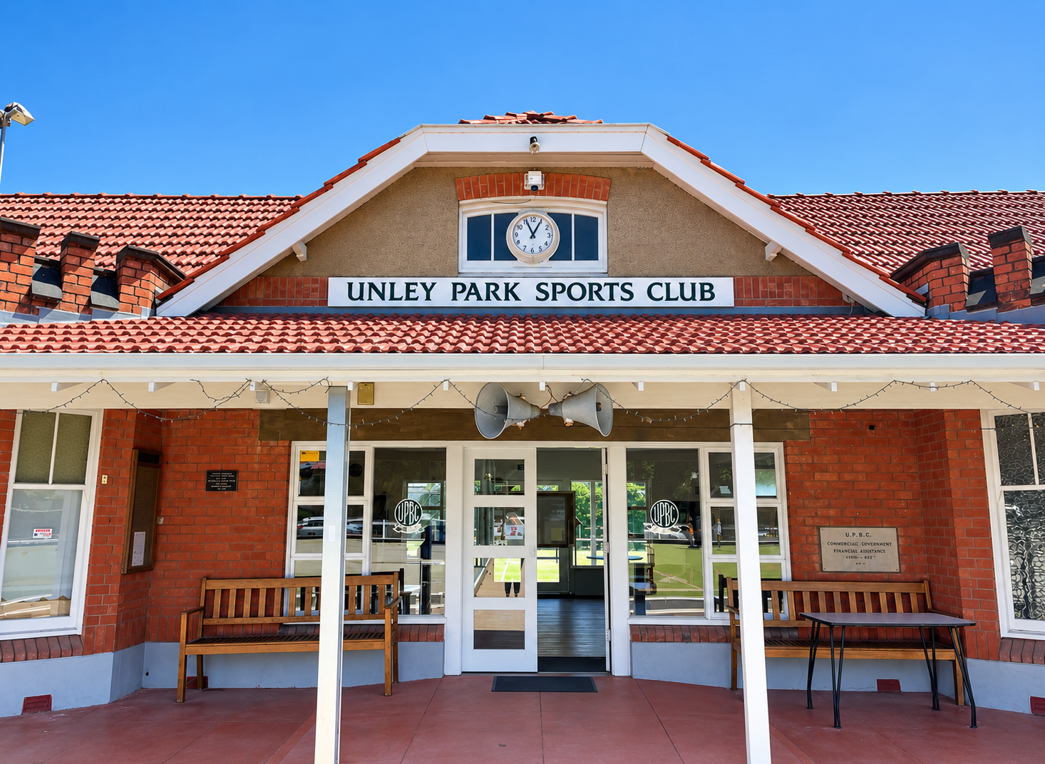 Front view of Unley Park Sports Club building with red tiled roof, two megaphones, a bench, and a sign indicating its name.