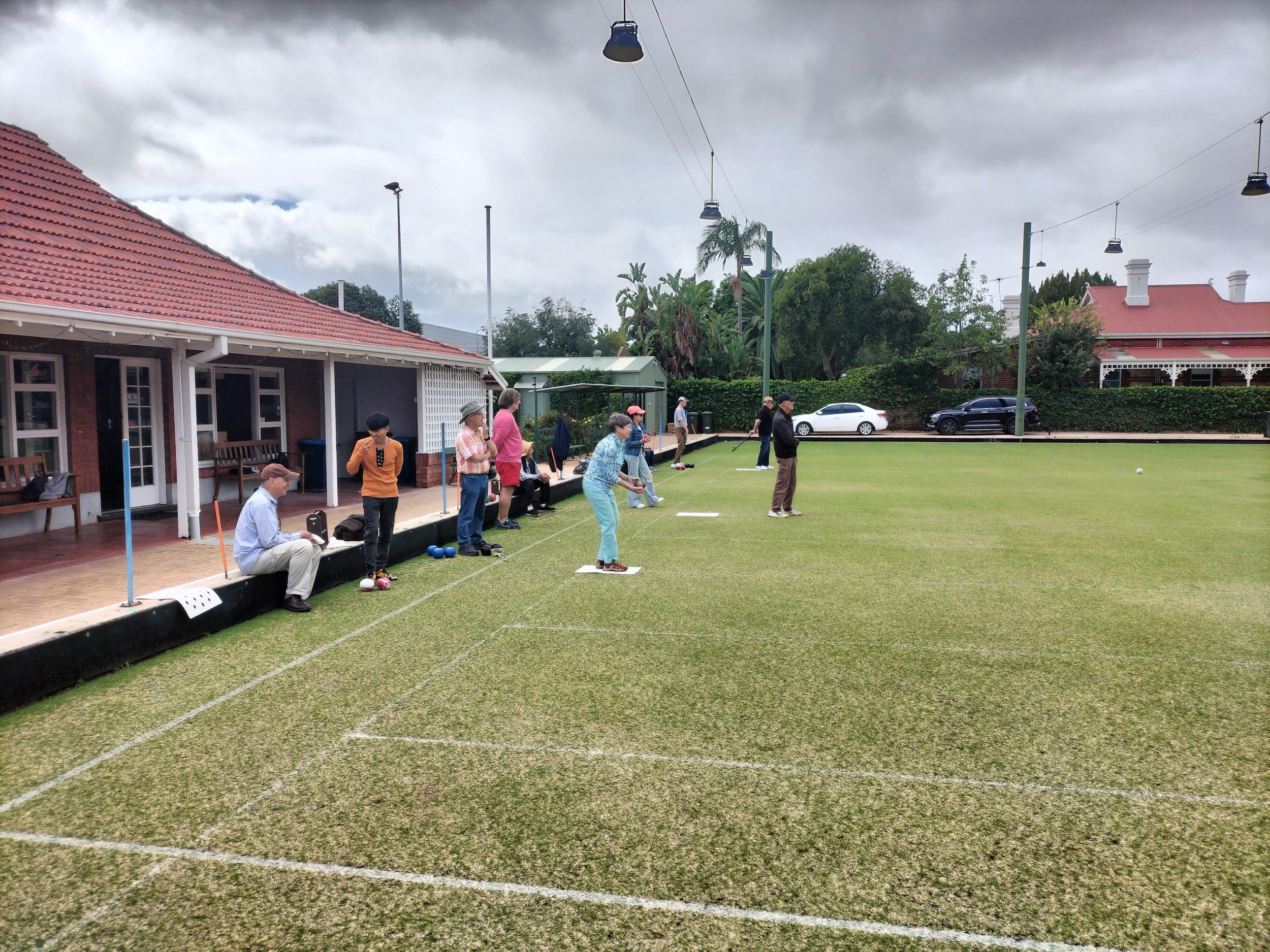 Several people are playing lawn bowls on a well-maintained grass field under a cloudy sky. Some are standing and some are leaning or sitting nearby, watching intently. There are buildings with red-tiled roofs and trees in the background.