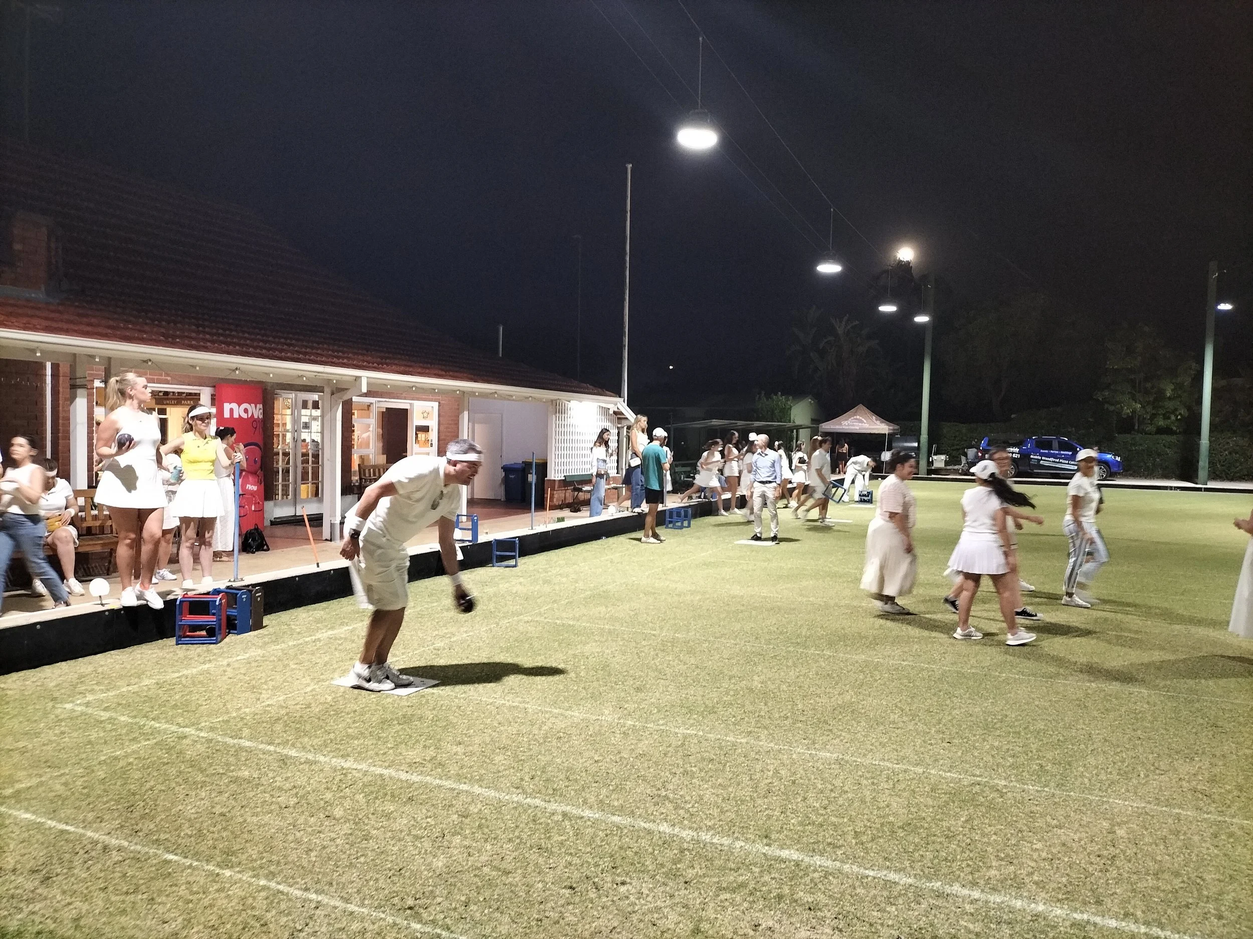 A group of people playing on a grass cricket field at night, with some individuals in white sportswear, a covered pavilion with people standing underneath, and floodlights illuminating the scene.