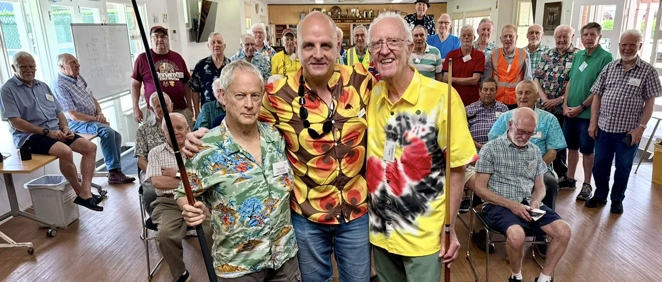 Group of elderly men and women gathered in a brightly lit room, some seated and some standing, with three men in the front wearing colorful Hawaiian shirts, one holding a cane. The background has large windows, a whiteboard, and a television.
