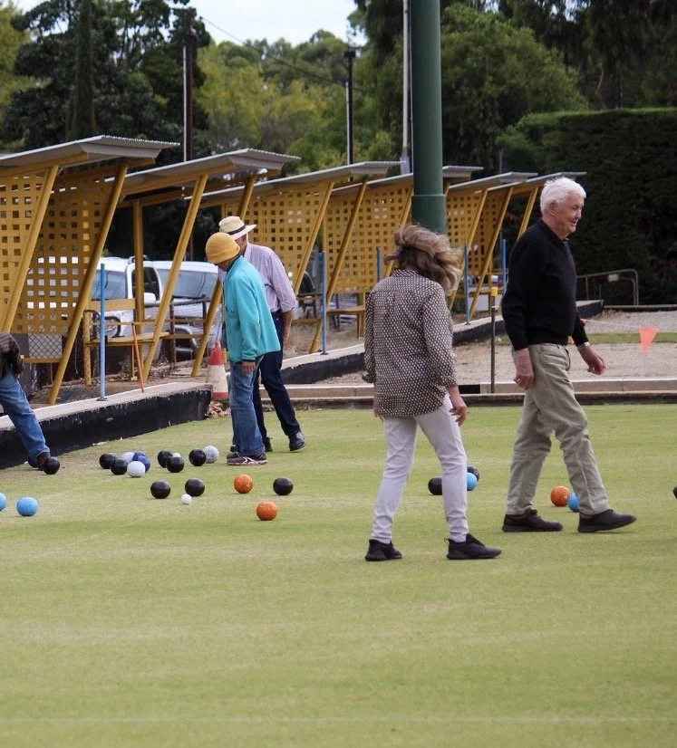 People playing lawn bowls on a green with wooden shelters in the background.