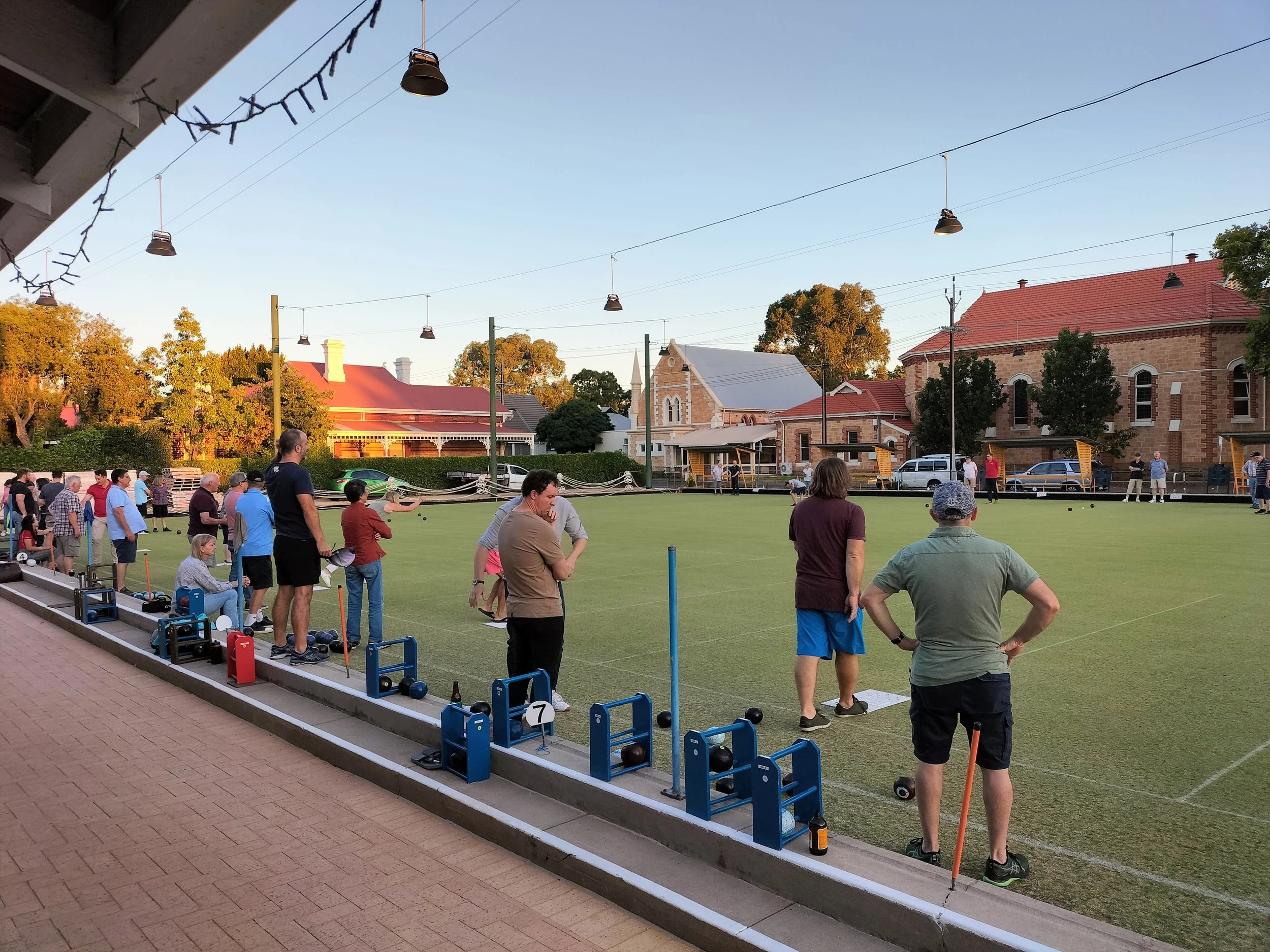 People playing lawn bowls on a green field with onlookers around in a small town setting, with strings of lights hanging overhead and historic buildings in the background.