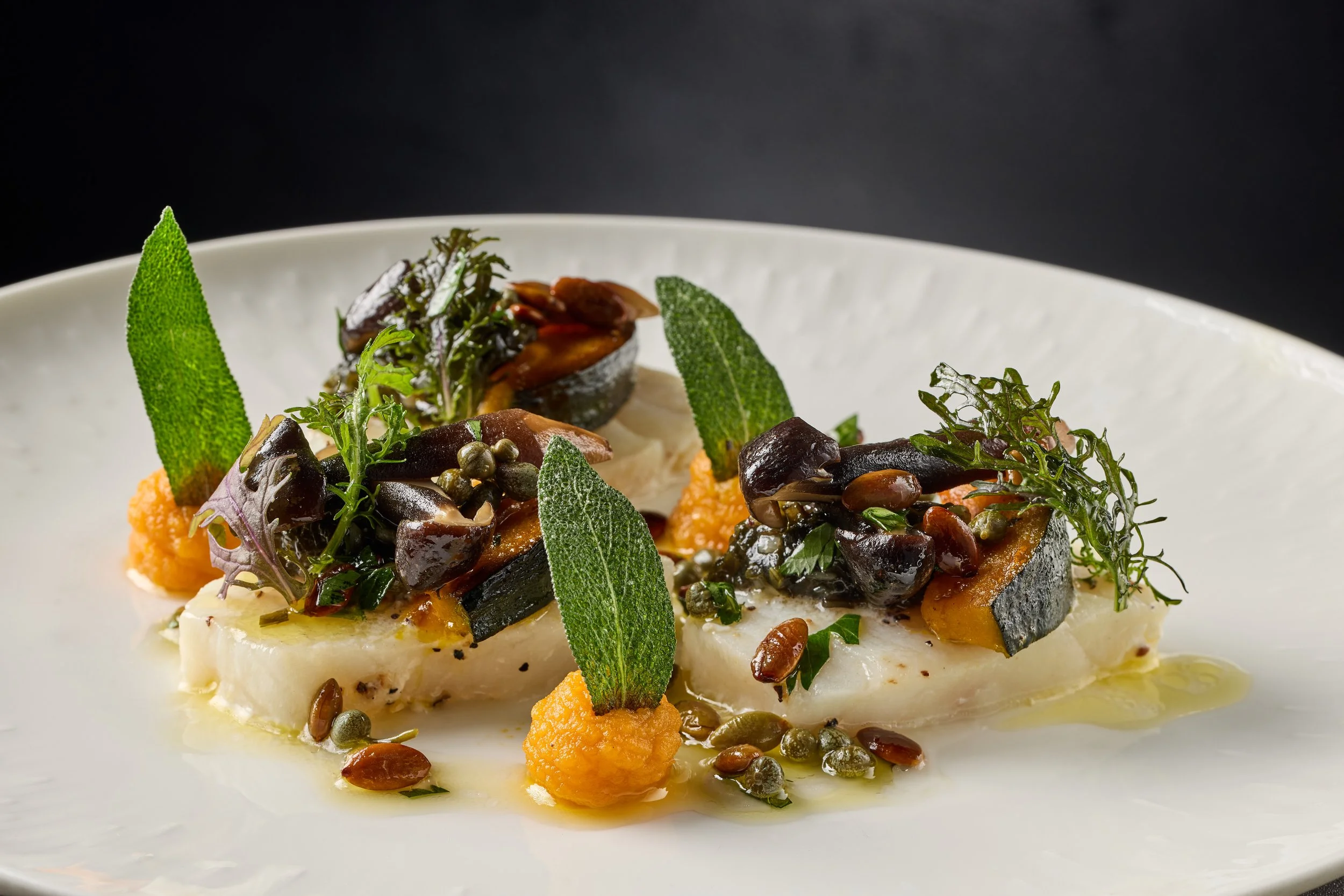 A gourmet dish featuring a base of white fish topped with herbs, mustard seeds, and grilled vegetables, garnished with sage leaves, served on a white plate against a black background.