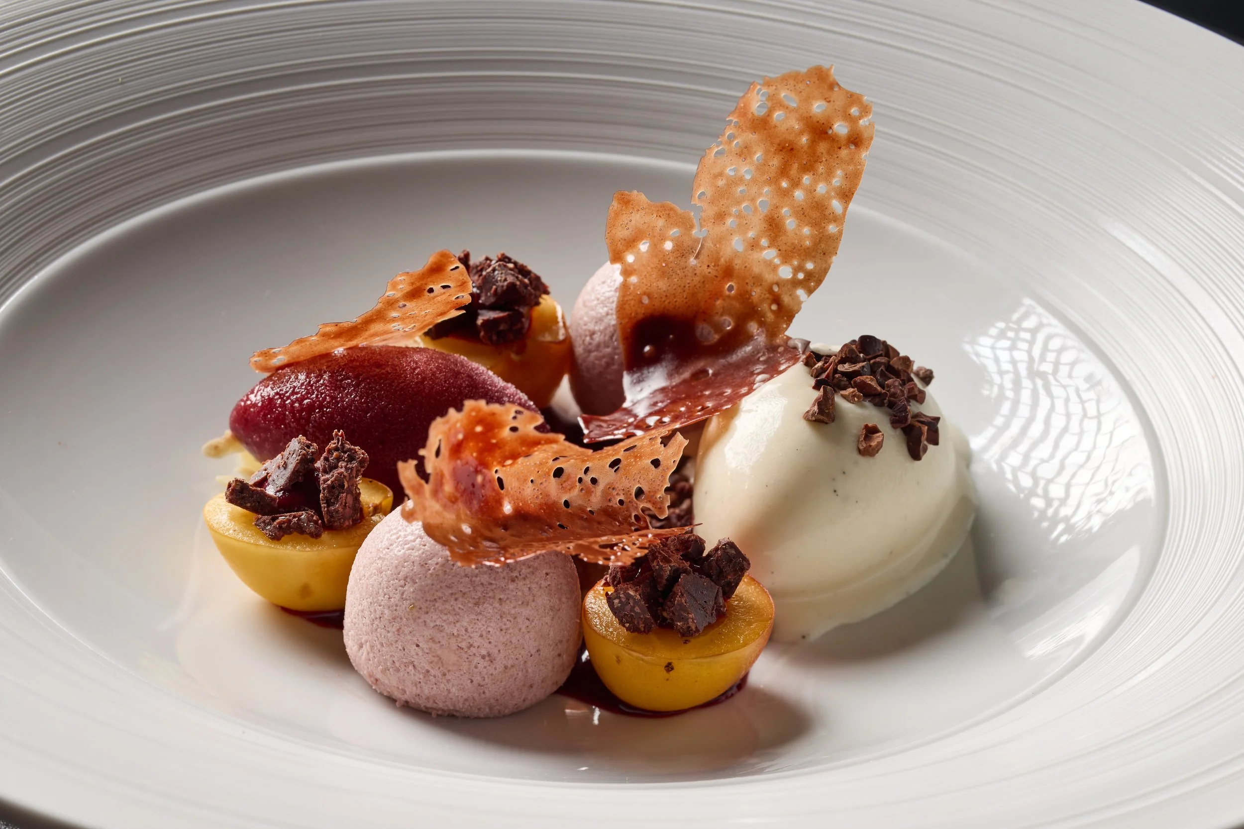 A plated dessert with purple and yellow fruit, pink sorbet, chocolate chunks, caramelized sugar and puffed rice.