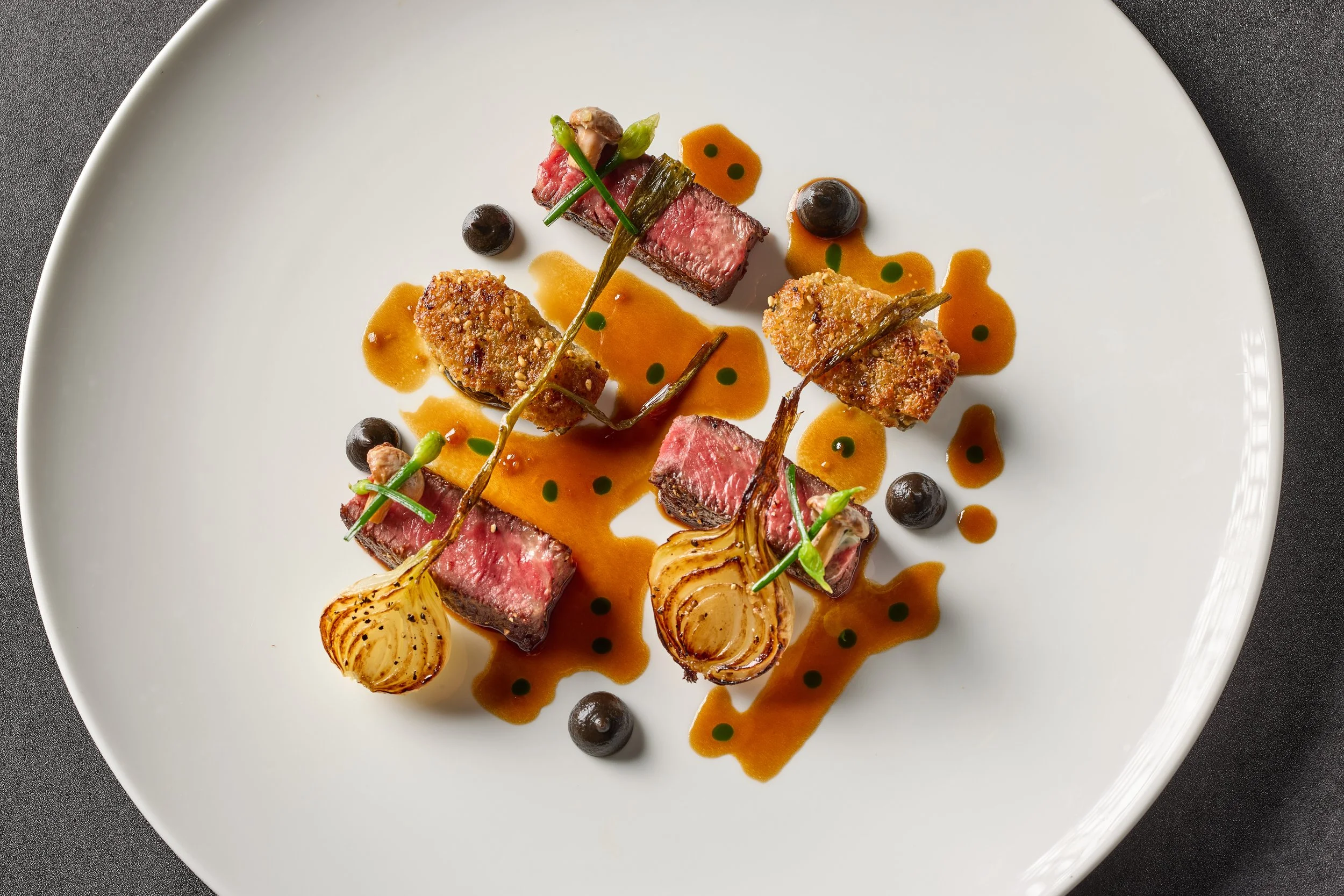 Seared steak sliced into pieces with sauce, grilled onions, black spheres (possibly berries), and garnishes on white plate.