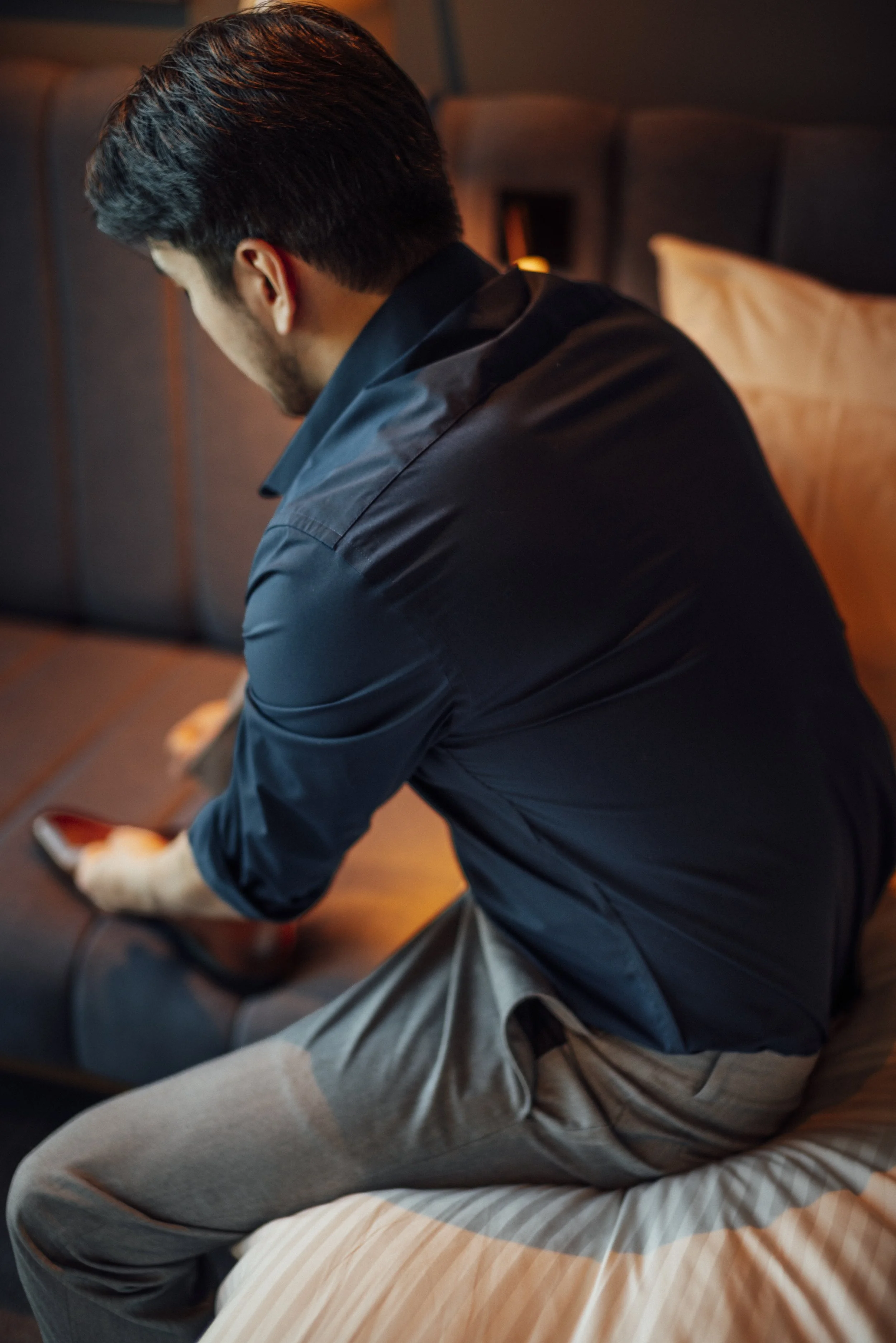 A man with dark hair wearing a dark shirt is sitting on a bed in a dimly lit room. The person is leaning forward, with their side profile visible, and appears to be looking down or engaged in an activity on the bed. The background includes a headboard and a pillow.