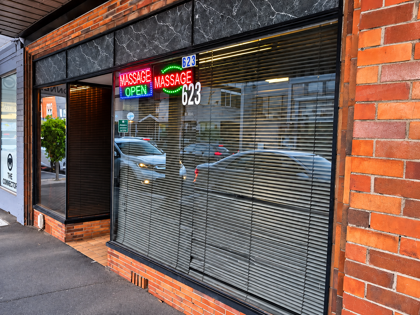 A storefront with neon signs reading 'Massage Open' in red, green, and blue, and the street view through closed blinds, showing parked cars and buildings.