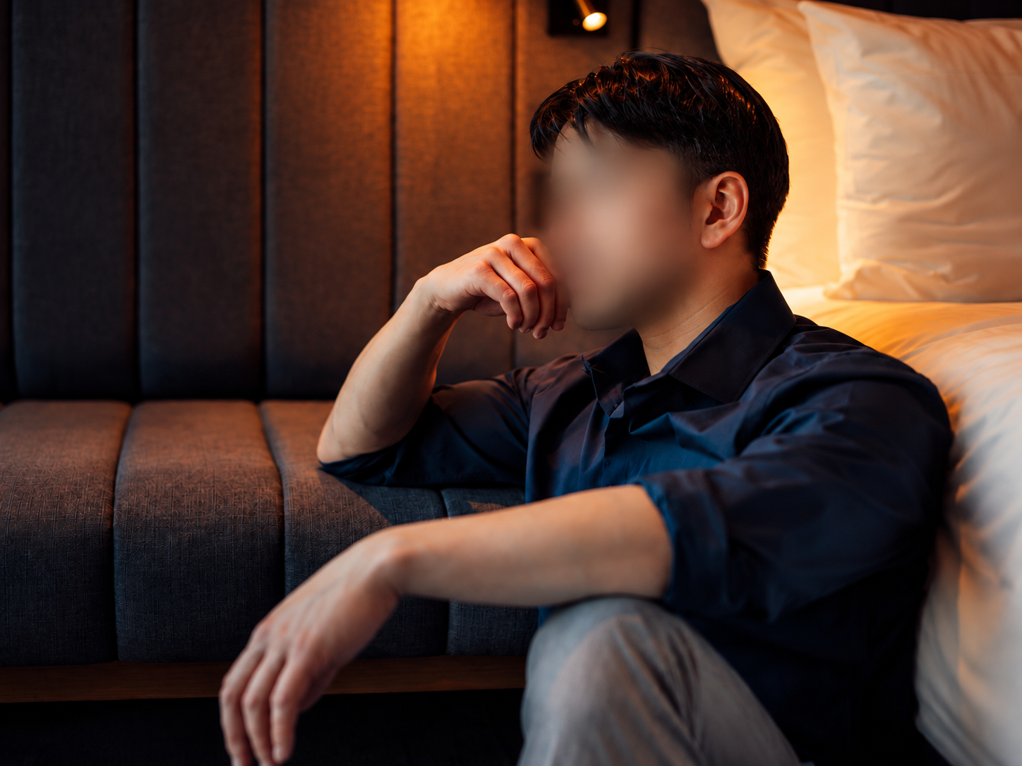 Man sitting on the floor beside a bed with a thoughtful expression, resting his face on his hand, in a dimly lit room.