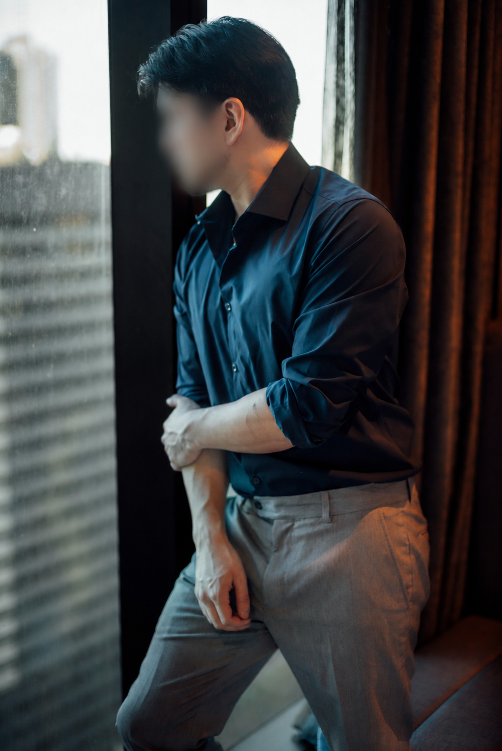 A man with dark hair dressed in a dark blue shirt and gray pants standing by a window, looking outside with a contemplative expression.