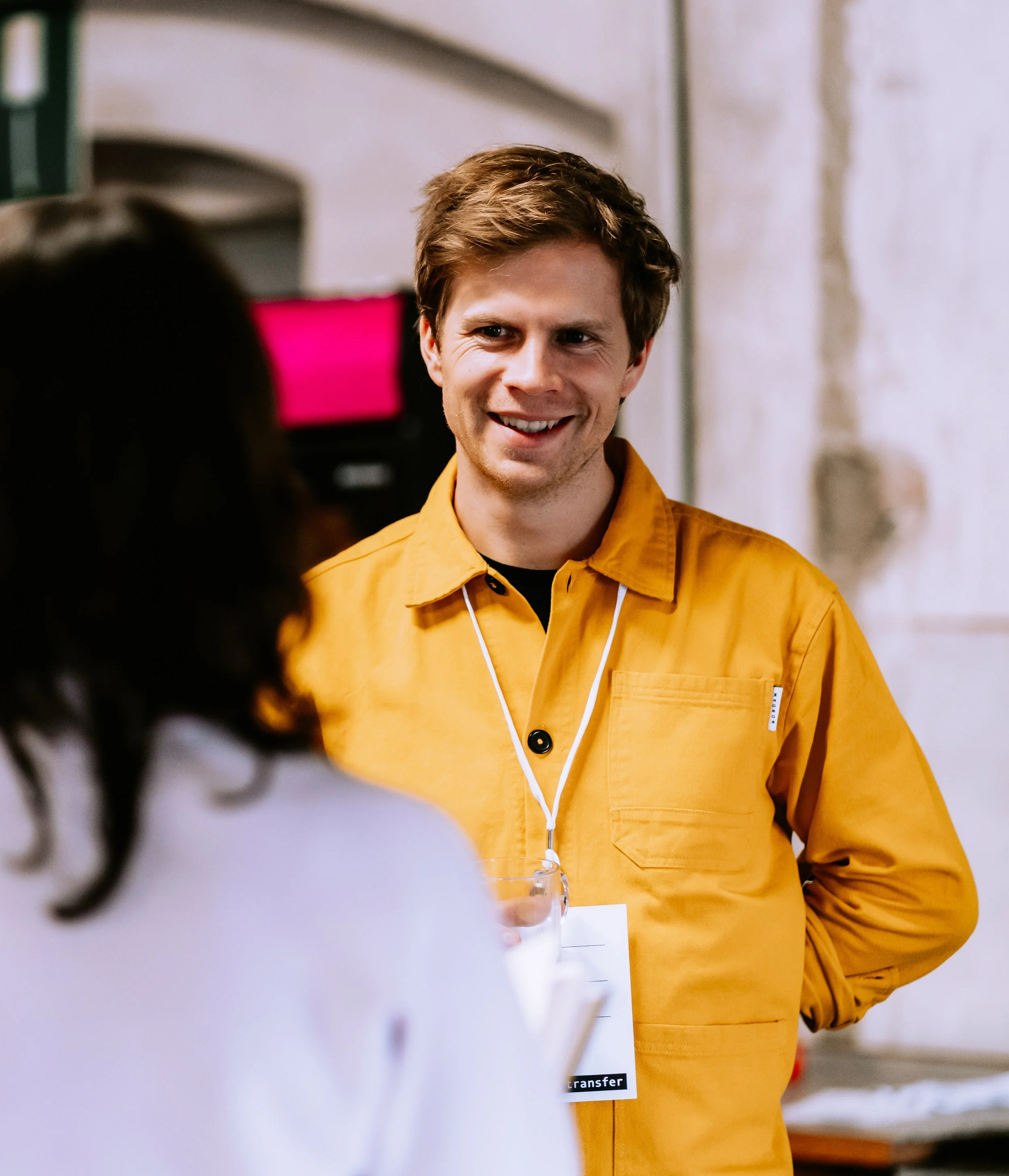 Ein lachender junger Mann in einem orangefarbenen Hemd wird interviewt, im Hintergrund ein weißer Innenraum.