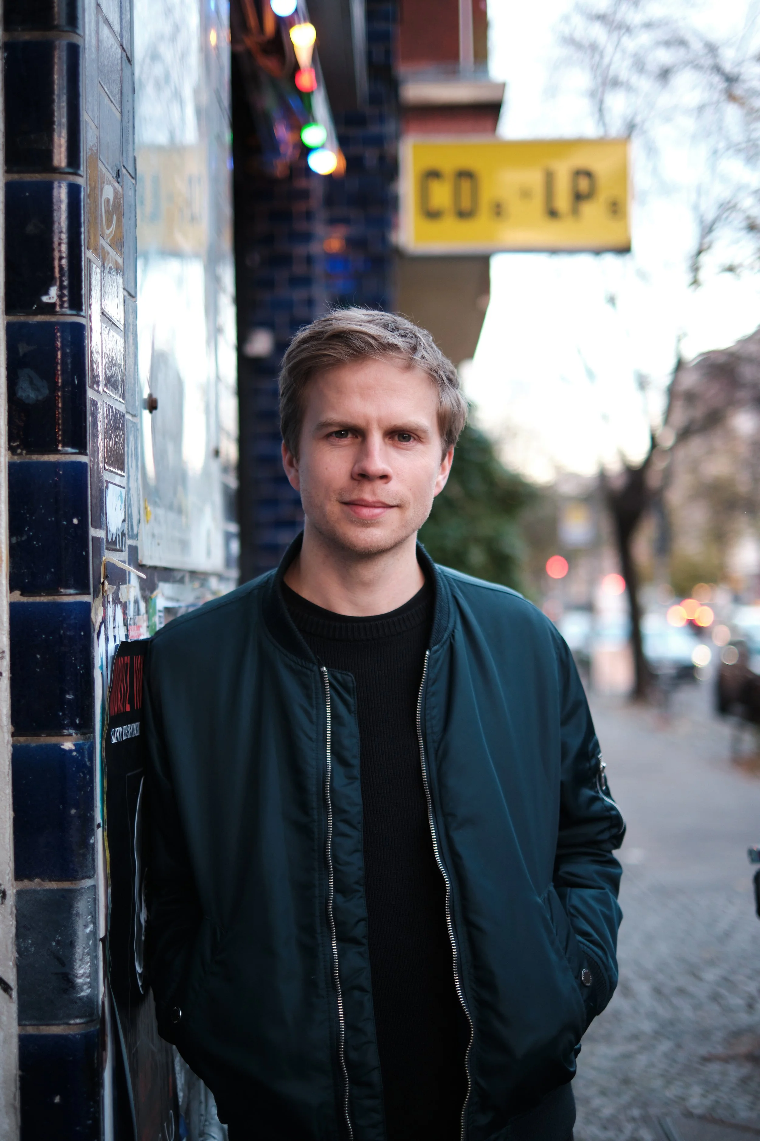 Junger Mann mit blondem Haar, dunkler Jacke mit Reißverschluss, steht an einer Wand mit Graffiti, im Hintergrund bunte Lichter und ein gelbes Schild mit der Aufschrift 'CO-LP', Bäume und eine Straße sind sichtbar.