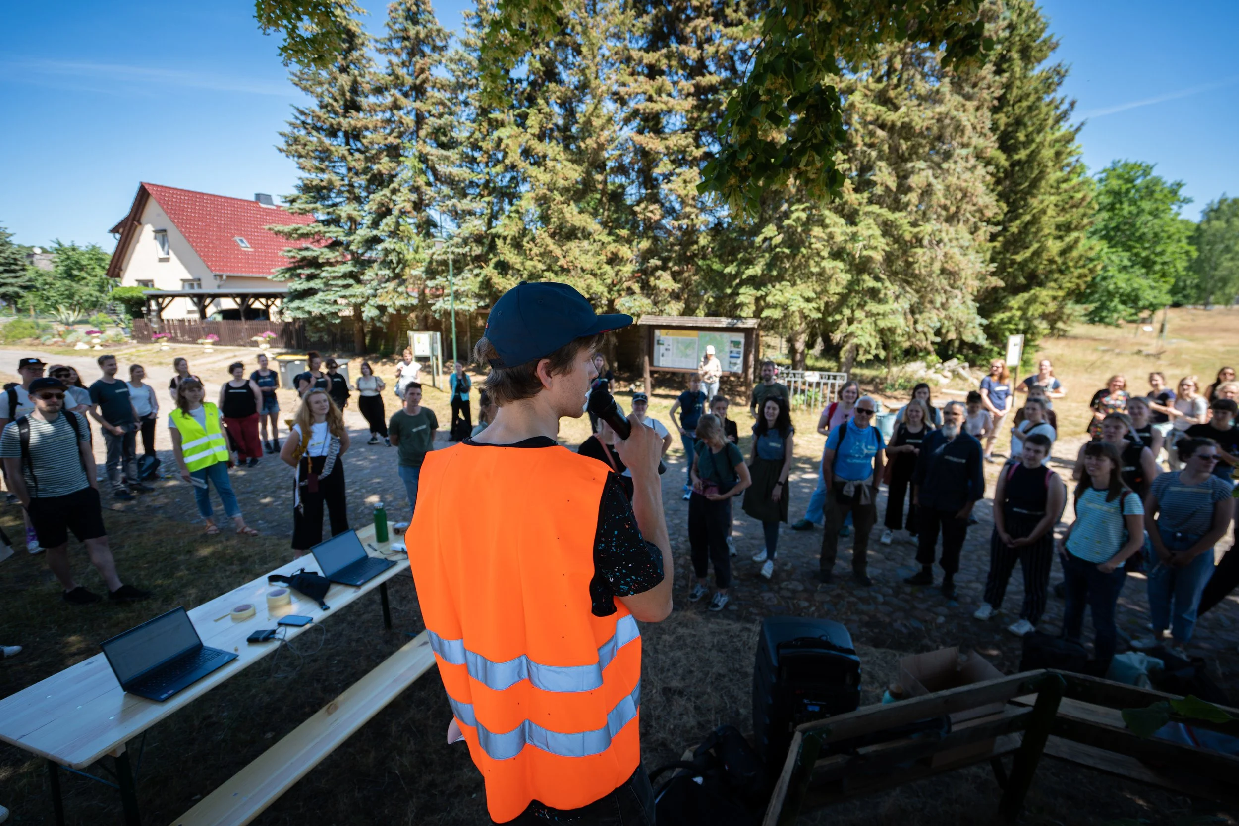 Eine Person in einem orangefarbenen Warnschutzwesten spricht auf einer Veranstaltung im Freien vor einer Gruppe von Menschen mit einem Mikrofon, hinter einem langem Tisch mit Laptops. Es ist sonnig, und im Hintergrund sind Bäume und ein Haus zu sehen.