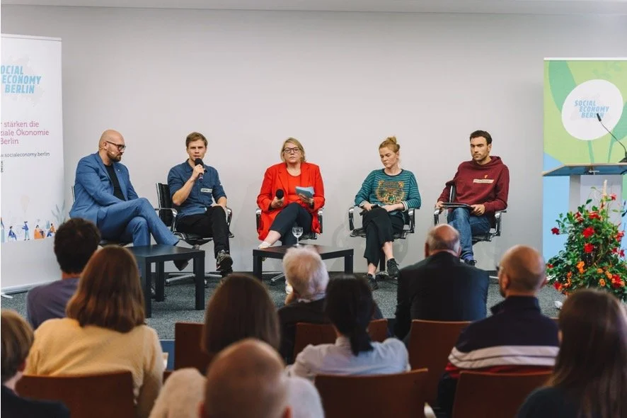 Fünf Personen sitzen auf einer Bühne während eines Panels bei einer Konferenz, mit einem Publikum im Vordergrund.