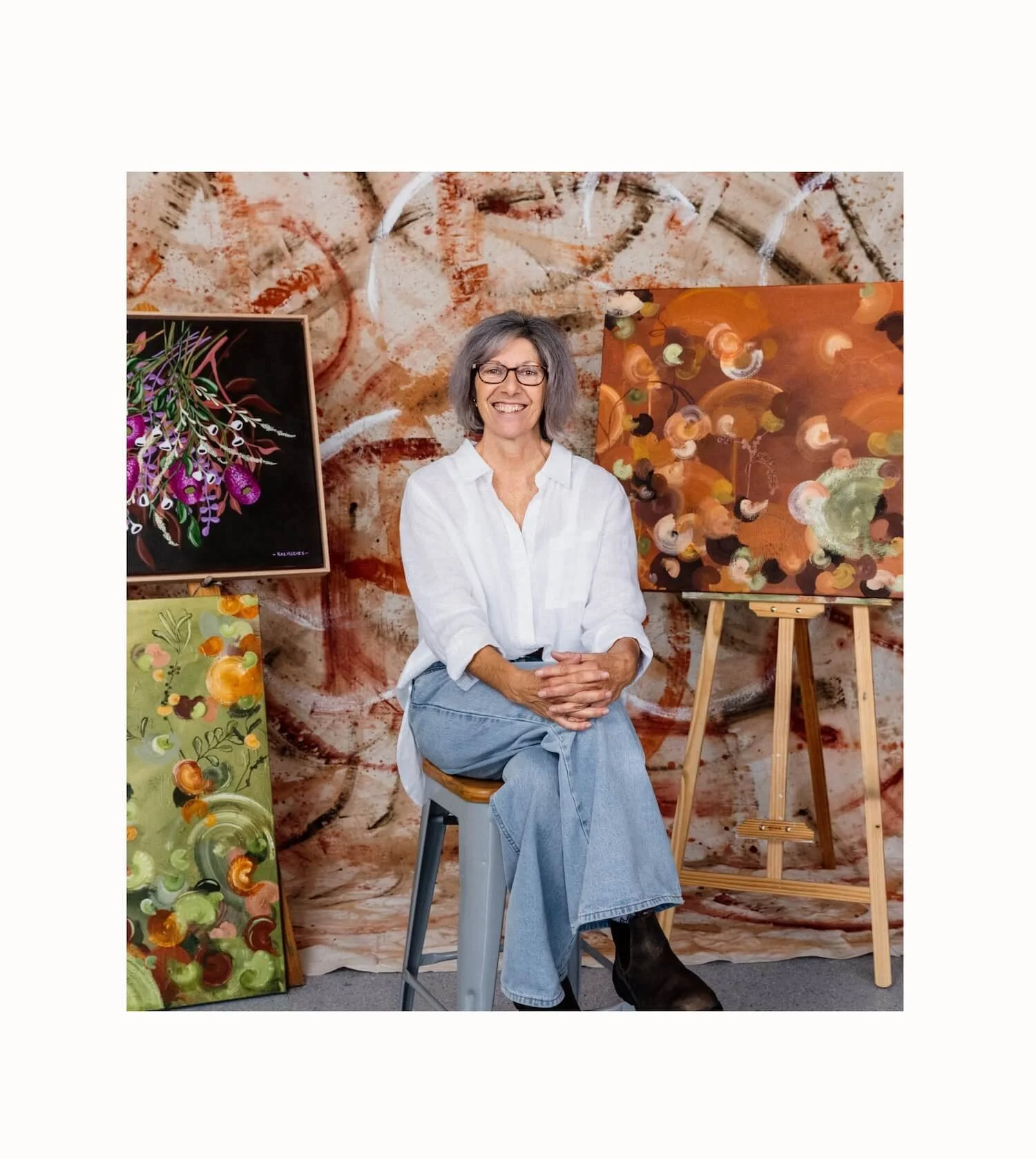 Artist Kaz Hughes sits on a chair in front of colorful abstract paintings in an art studio.