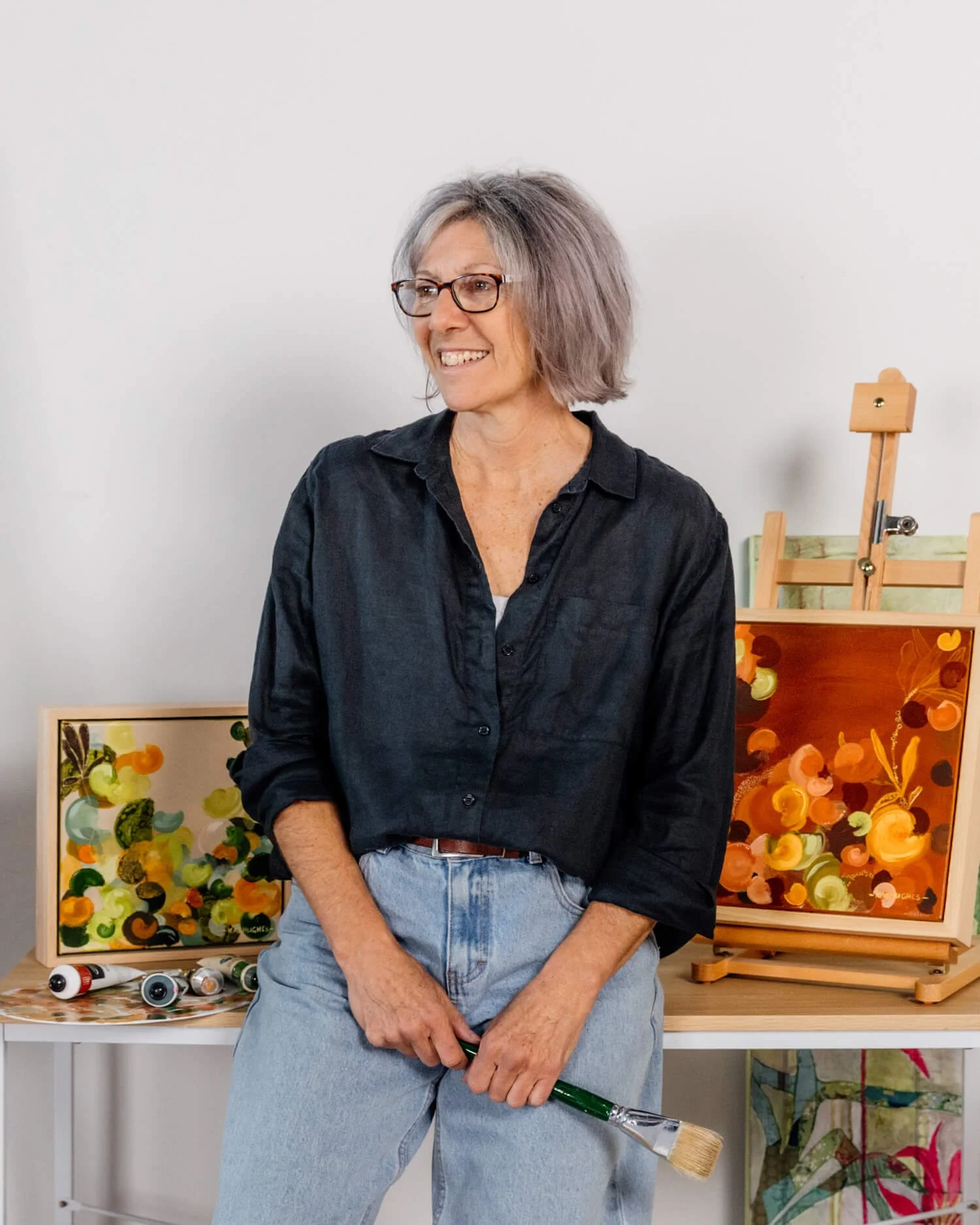 Artist Kaz Hughes, holding a paintbrush, standing in front of abstract paintings.