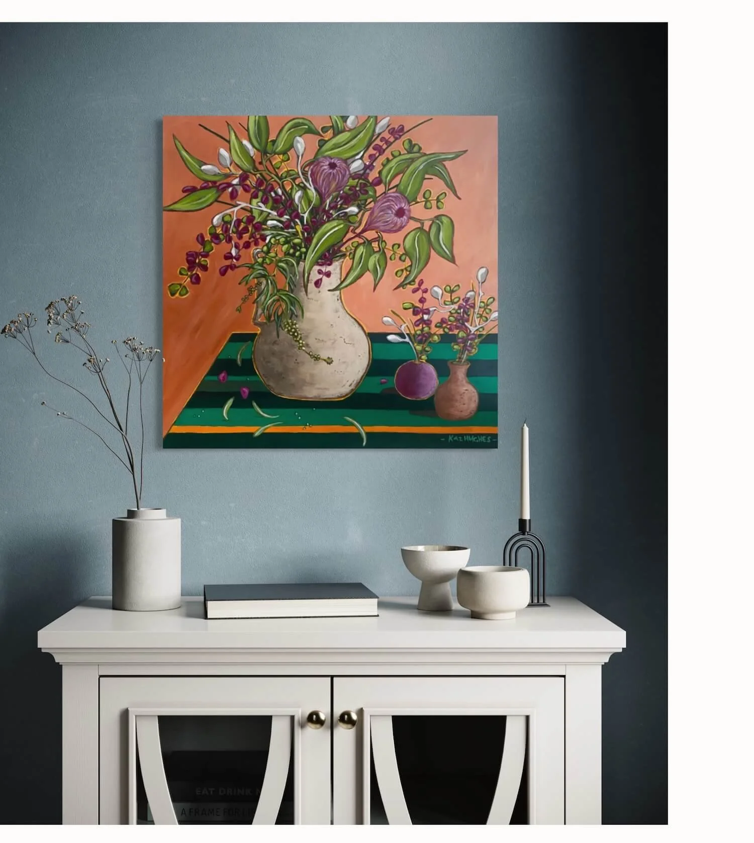 Interior of a modern room with a white console table decorated with a vase of dried flowers, bowls, a candle, and a decorative black candle holder. A colorful floral painting hangs on a teal wall above the table.