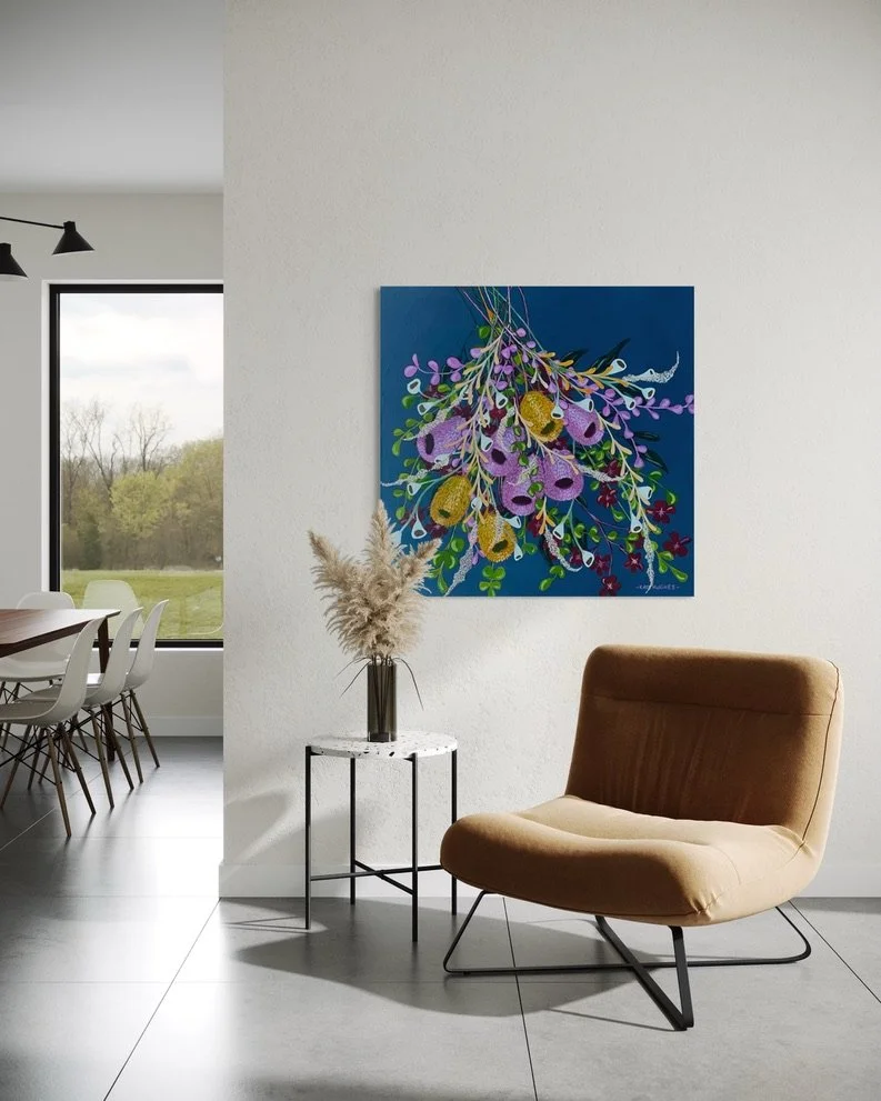 Modern living room corner with a beige armchair, a small marble side table with a vase and pampas grass, and a colorful floral painting on a white wall. Dining area with white chairs and a window showing an outdoor view of trees.