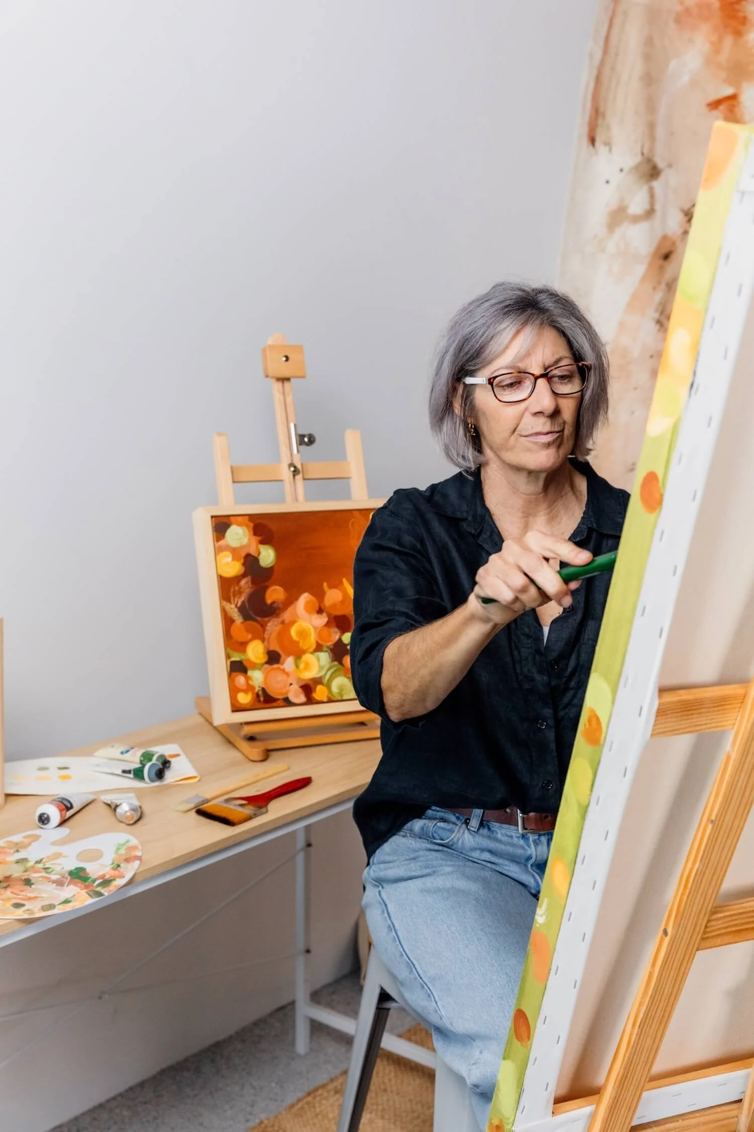 Artist, Kaz Hughes is painting on a canvas set on an easel. She is holding a green paintbrush and appears focused on her artwork.