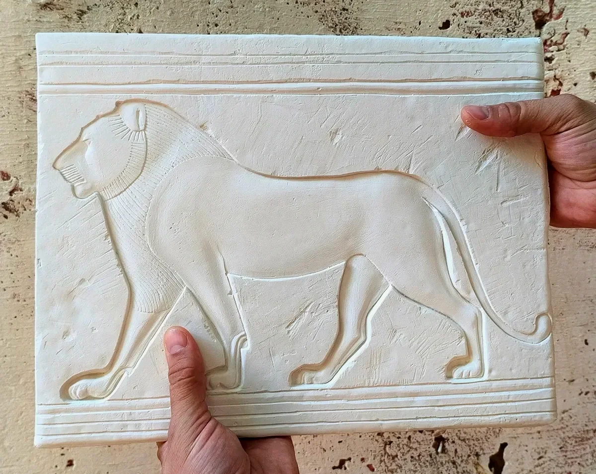 A person holding a carved white stone plaque featuring a raised outline of a lion in profile, with a detailed mane and tail, against a sandy background.