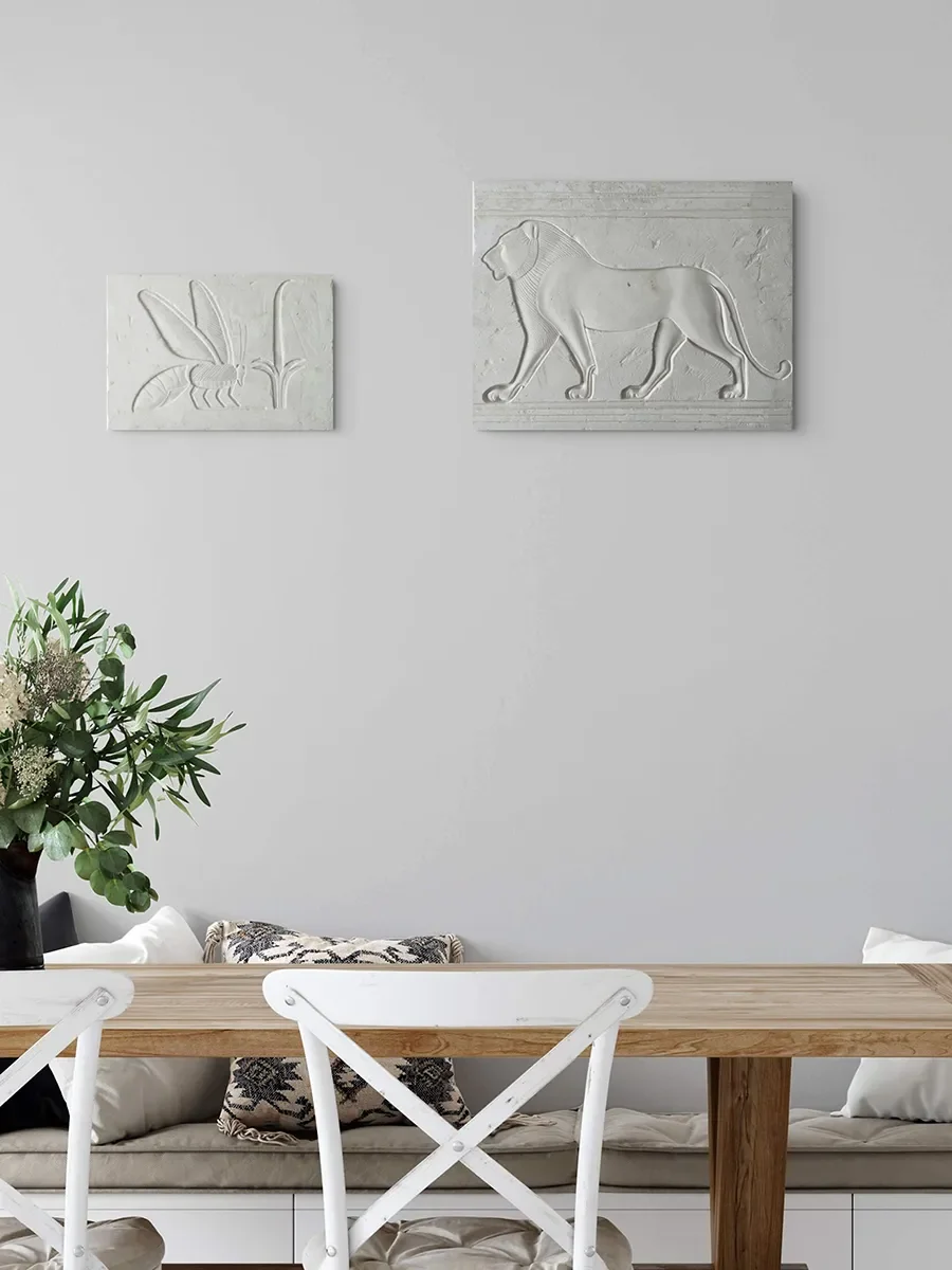 A minimalist dining area with a wooden table, white chairs, and wall art including a carved lion and a plant in a black vase, ancient art at home