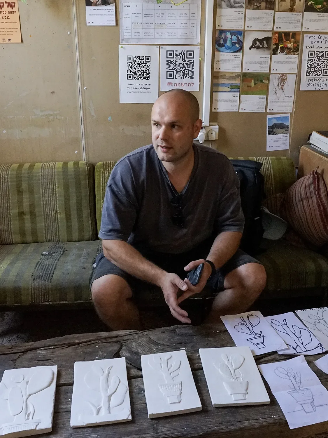 A bald man sitting on a green striped couch in an indoor setting with a bulletin board behind him that has various notices, QR codes, and photos. He is holding a smartphone in his right hand and there are drawings of potted plants on paper and foam in front of him on a wooden table.