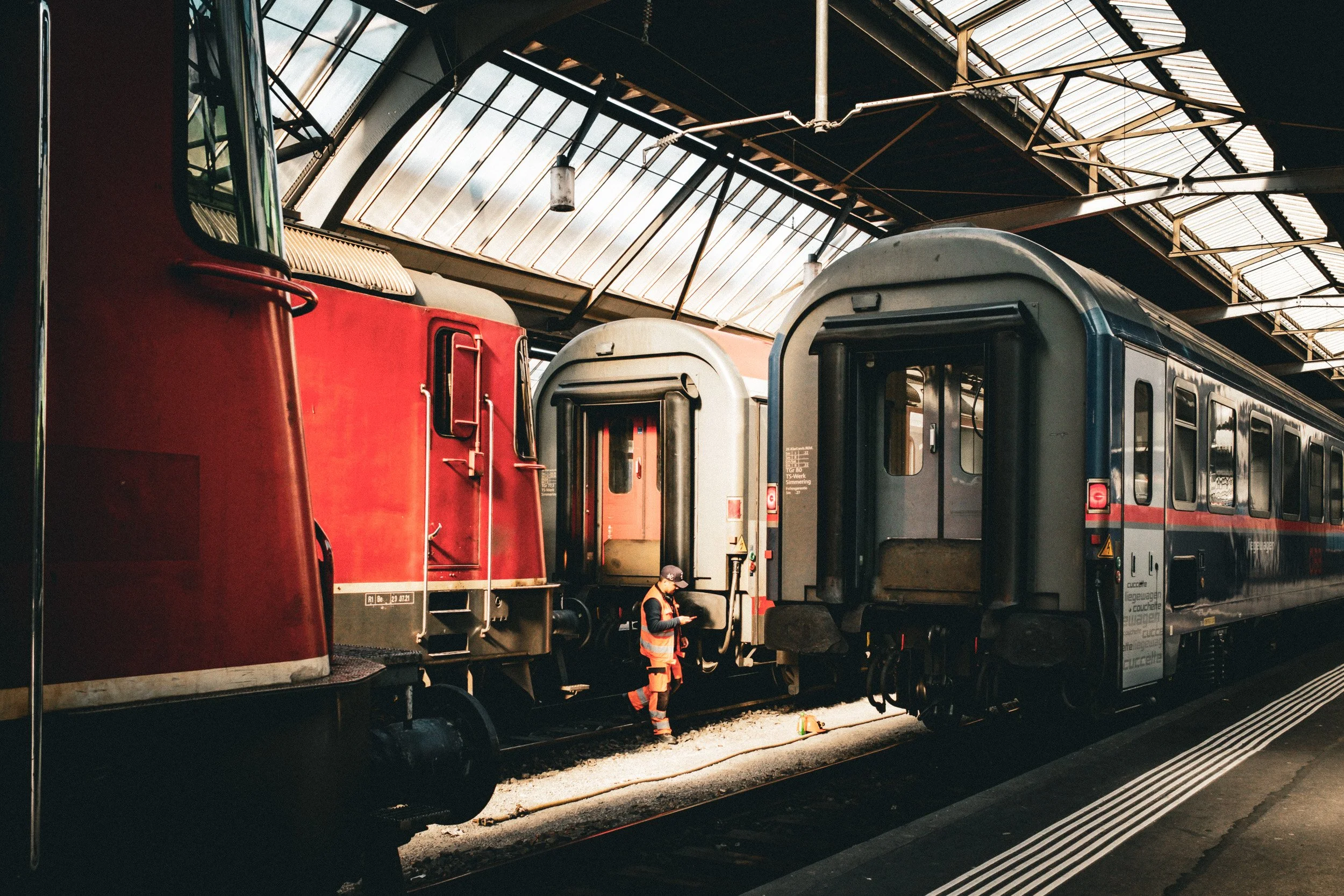 Railway worker stands between red and silver trains under a sunlit station roof.