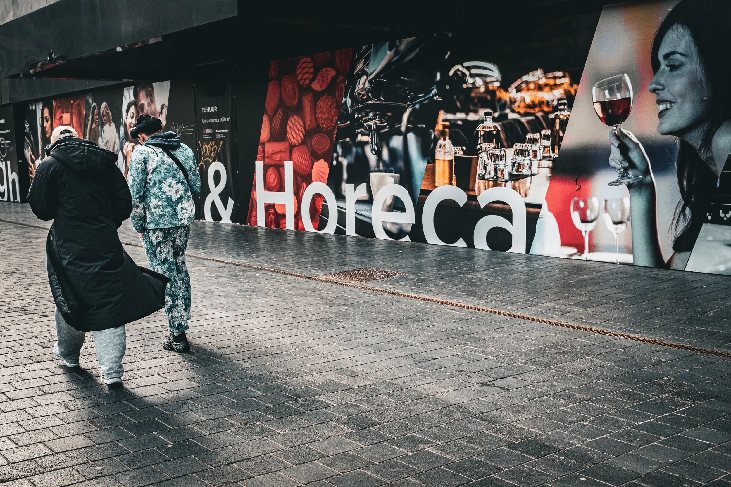 Two pedestrians walk beside a large advertisement showing a smiling woman holding a glass of wine.